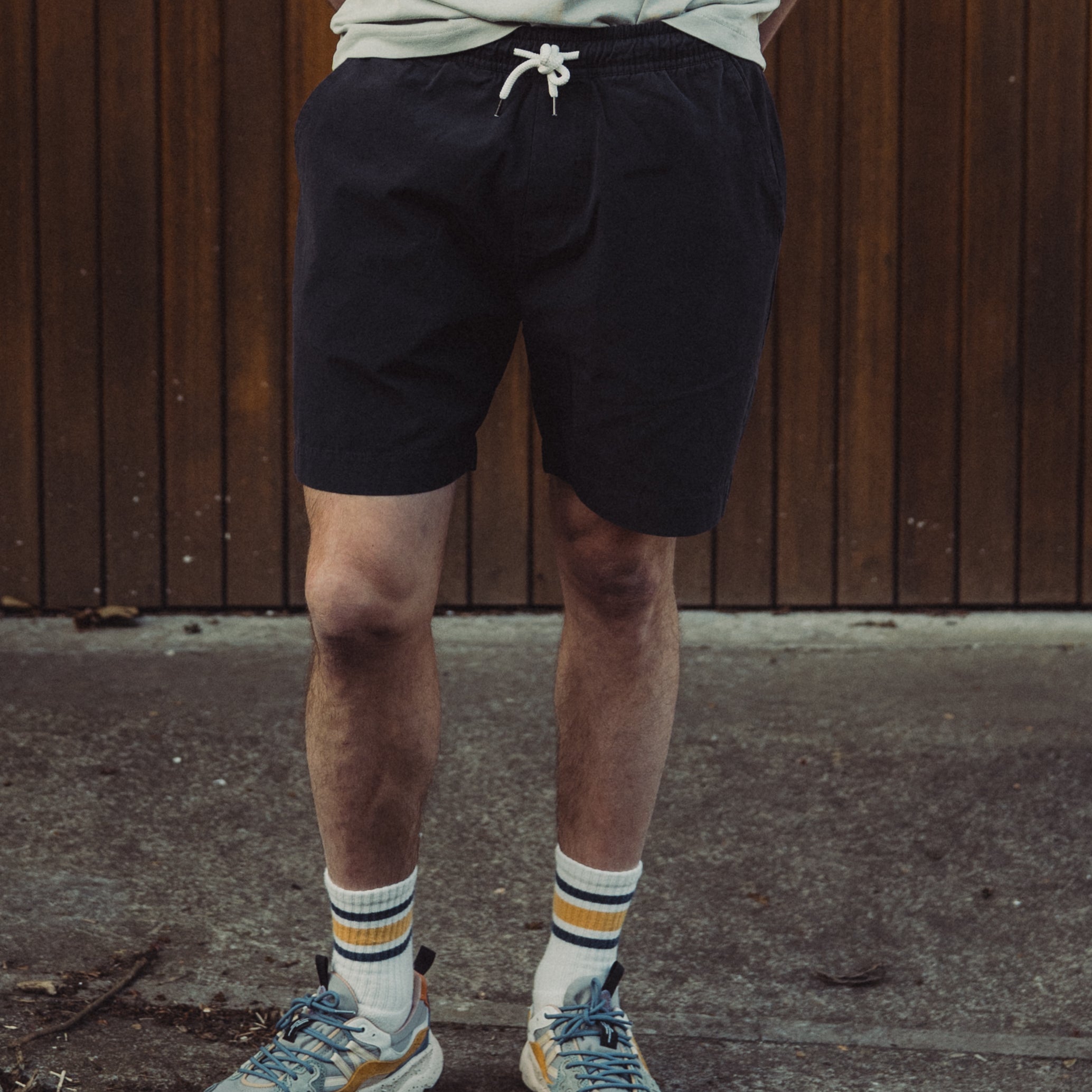 Man standing against a wooden wall wearing a light green t-shirt, black shorts, and colorful sneakers.