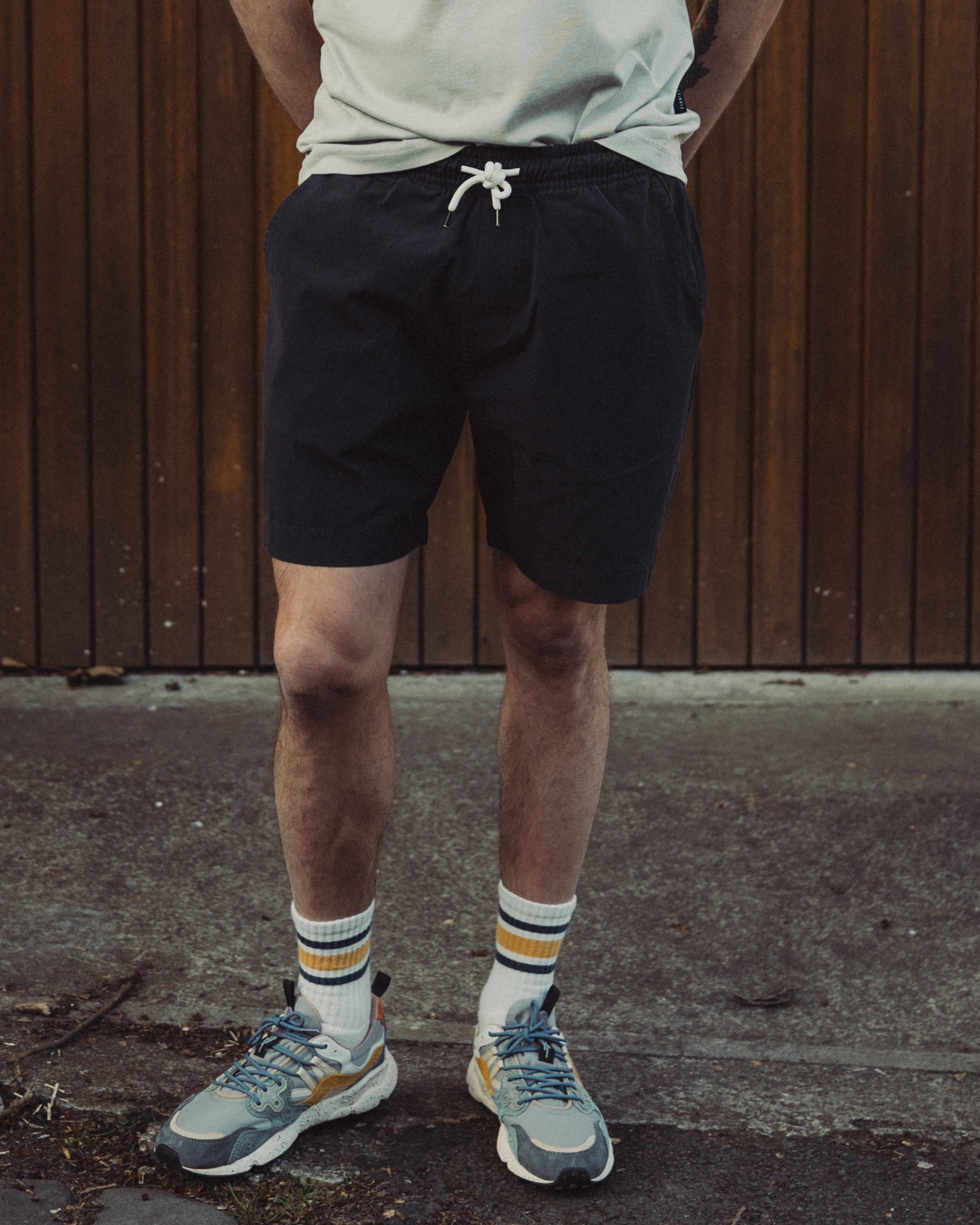 Man standing against a wooden wall wearing a light green t-shirt, black shorts, and colorful sneakers.
