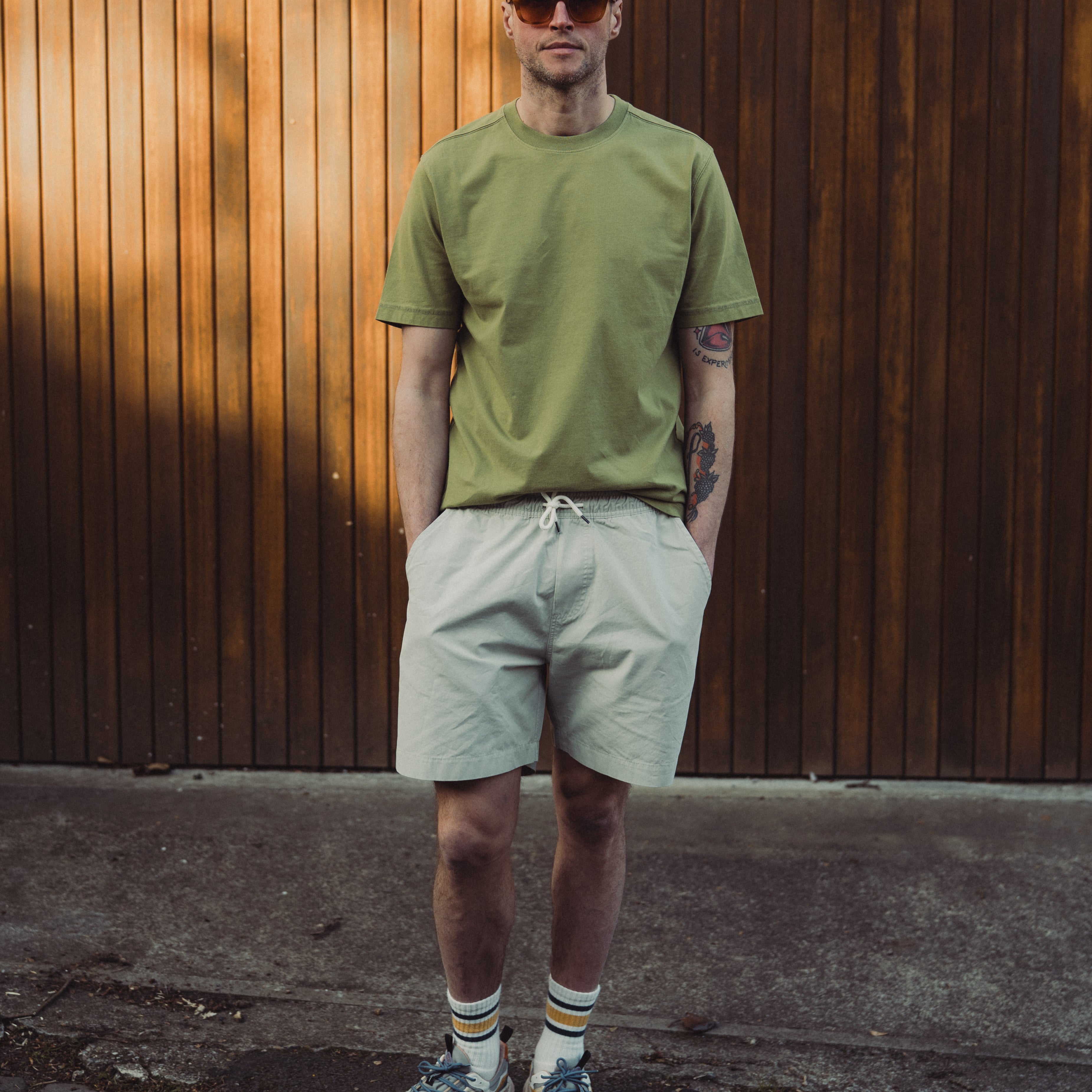 Man wearing a green t-shirt and light-colored shorts standing against a wooden wall.