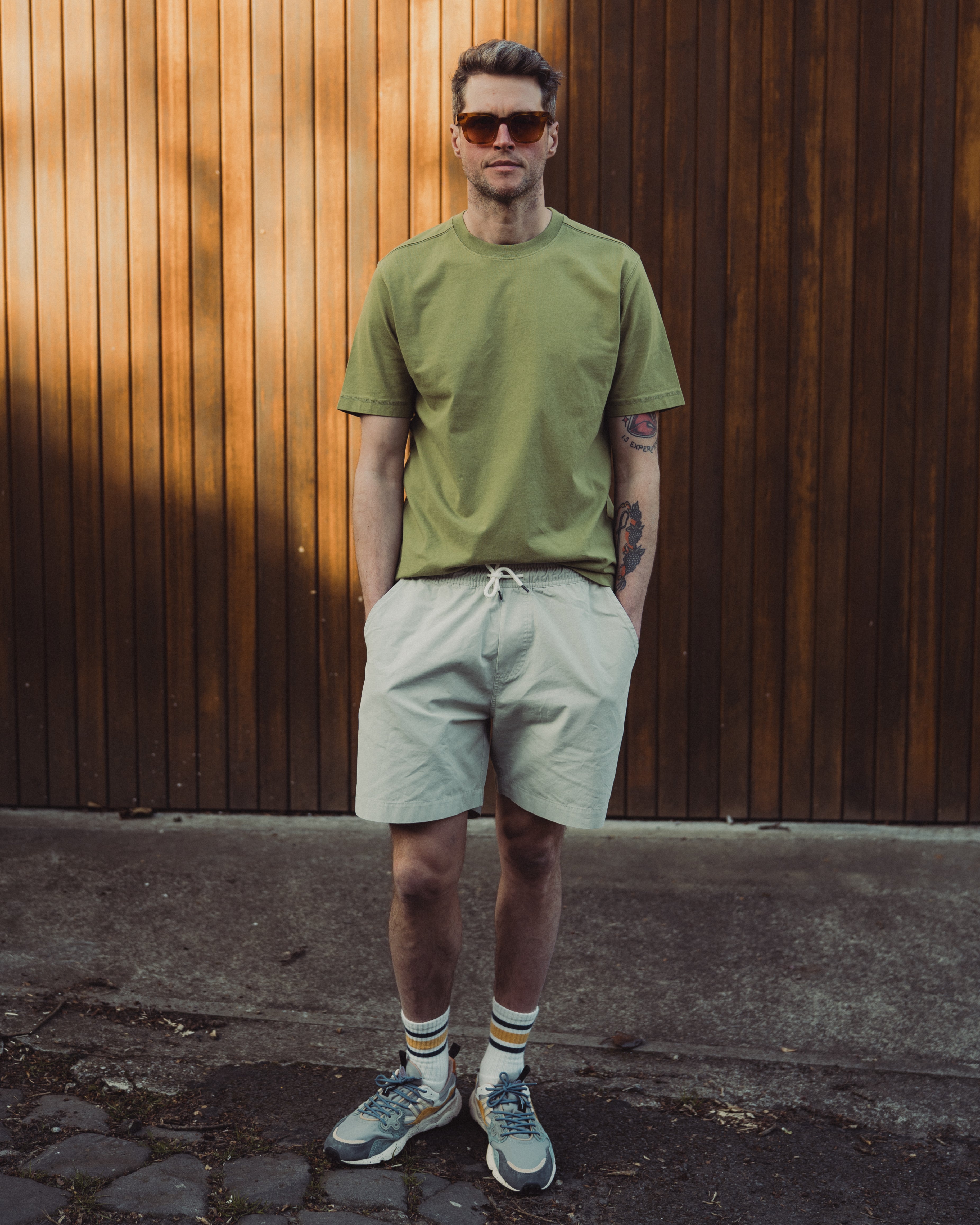 Man wearing a green t-shirt and light-colored shorts standing against a wooden wall.
