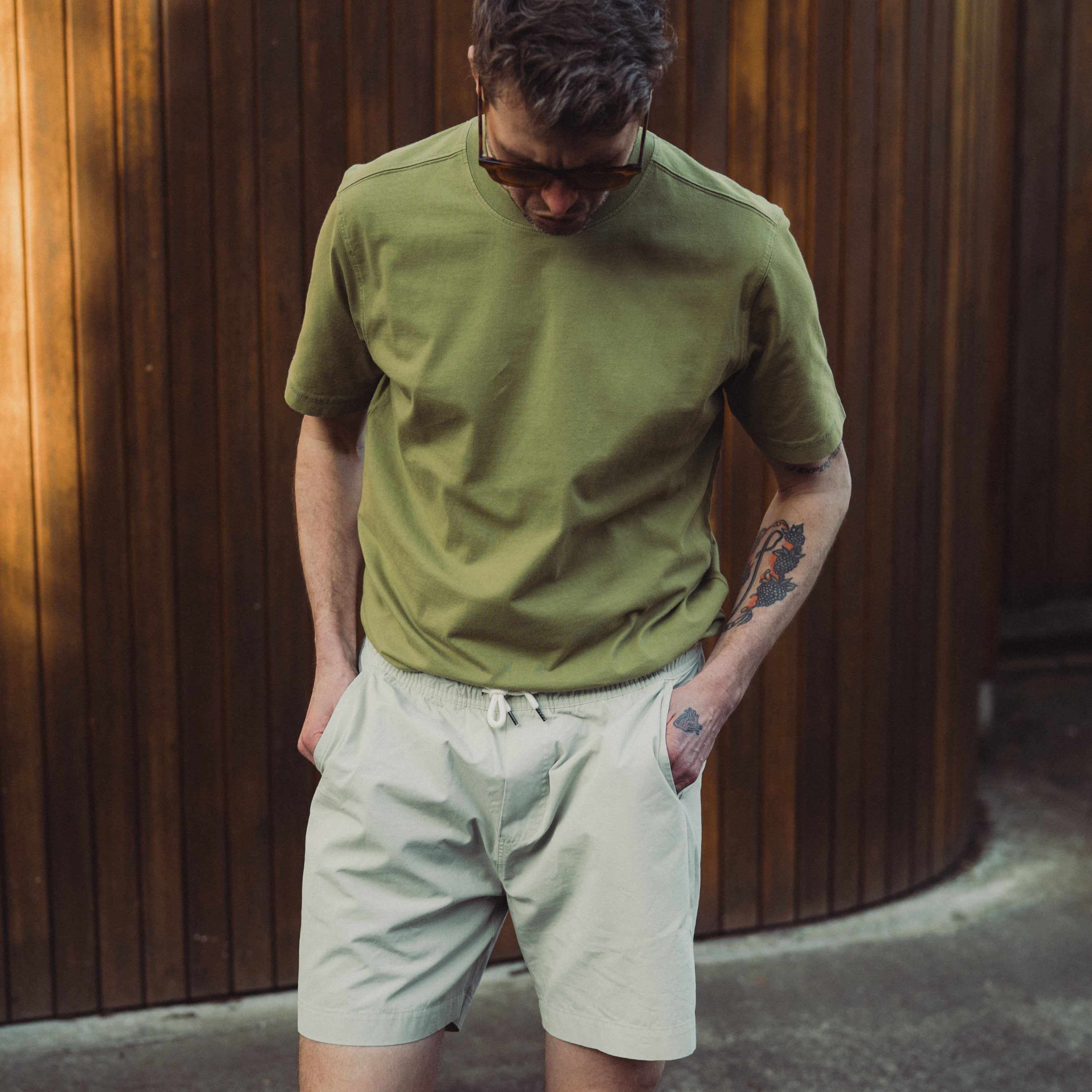 Man wearing a green shirt and beige shorts against a wooden wall.