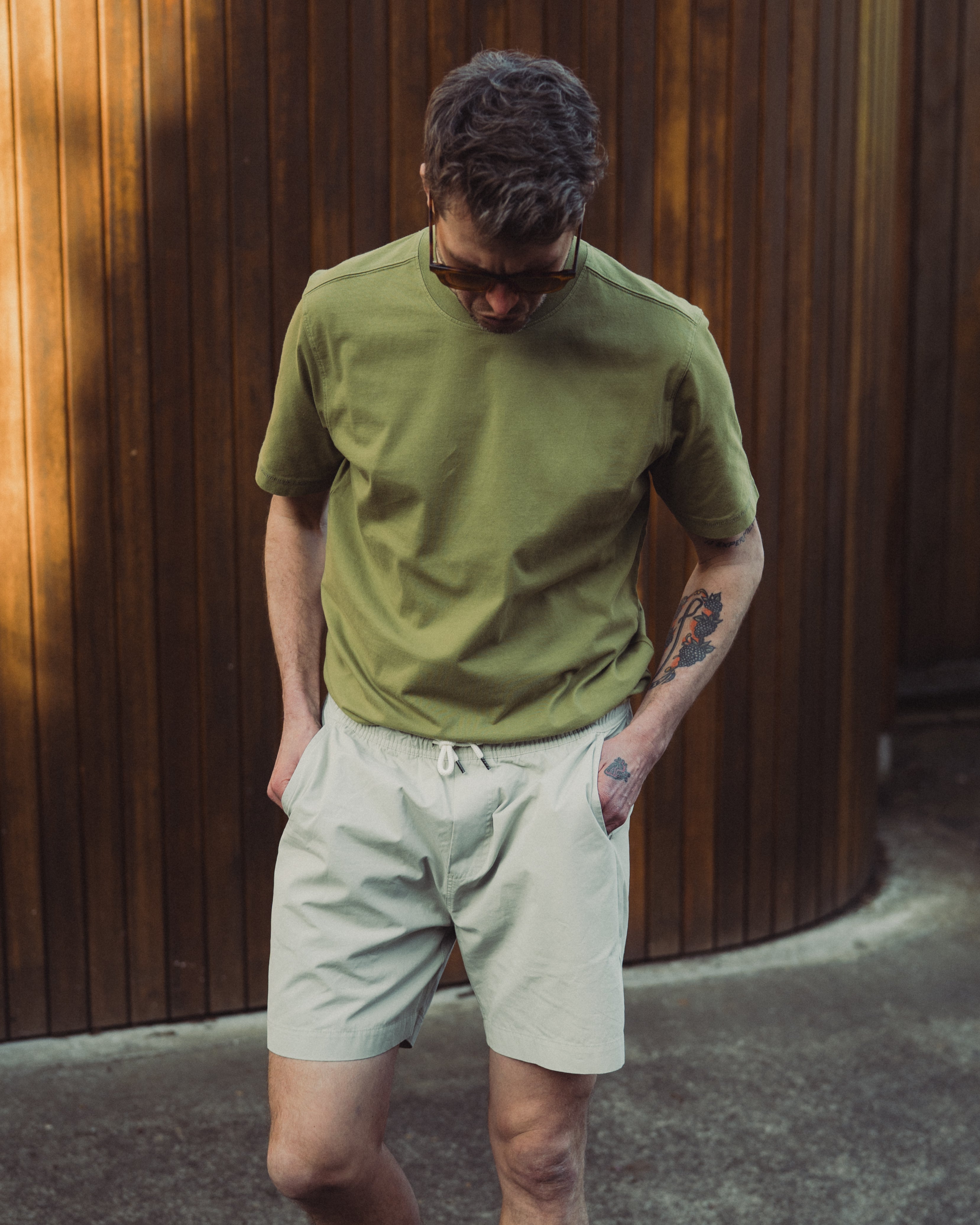 Man wearing a green shirt and beige shorts against a wooden wall.