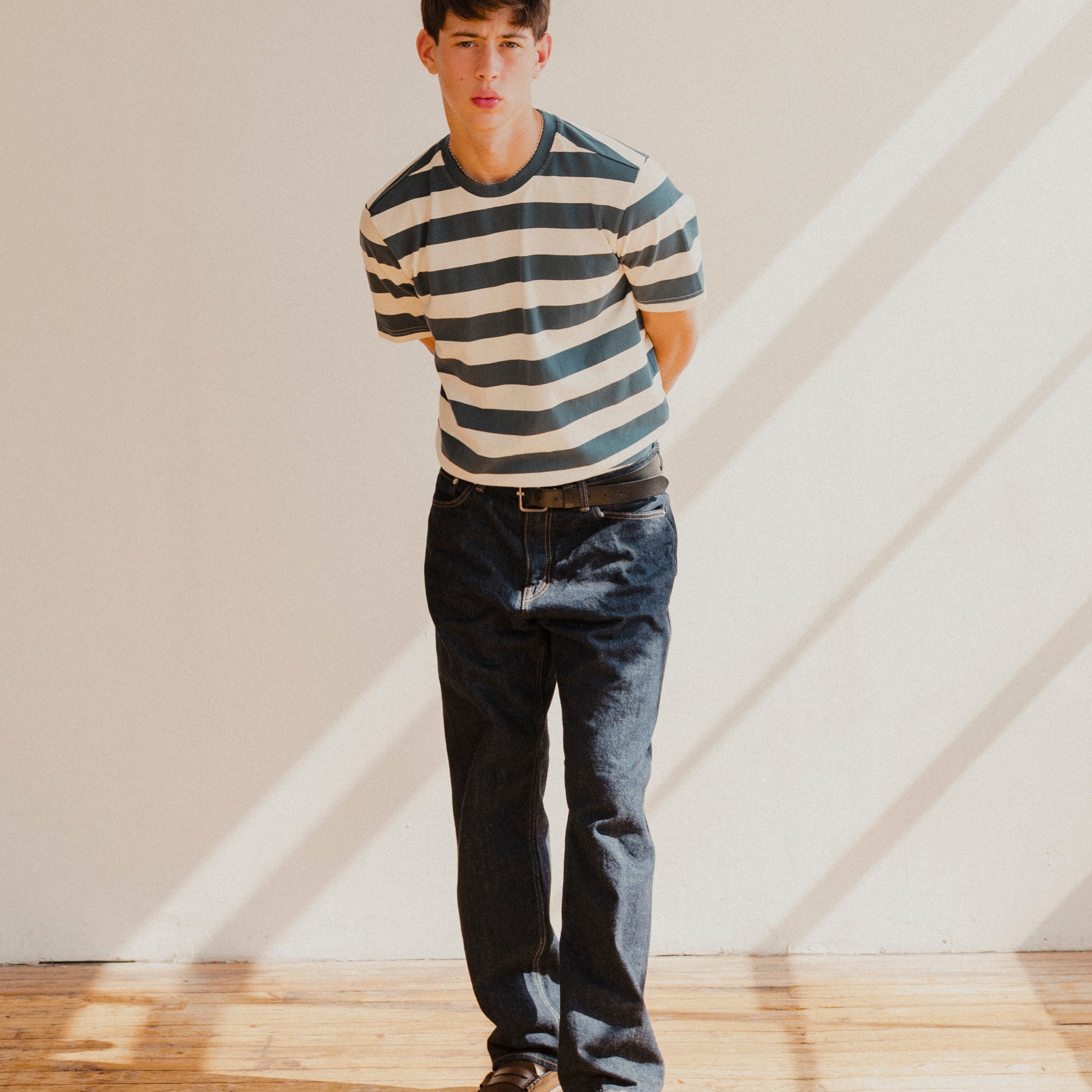 Man wearing a blue and white striped t-shirt and jeans standing in a room with wooden floor and white walls.