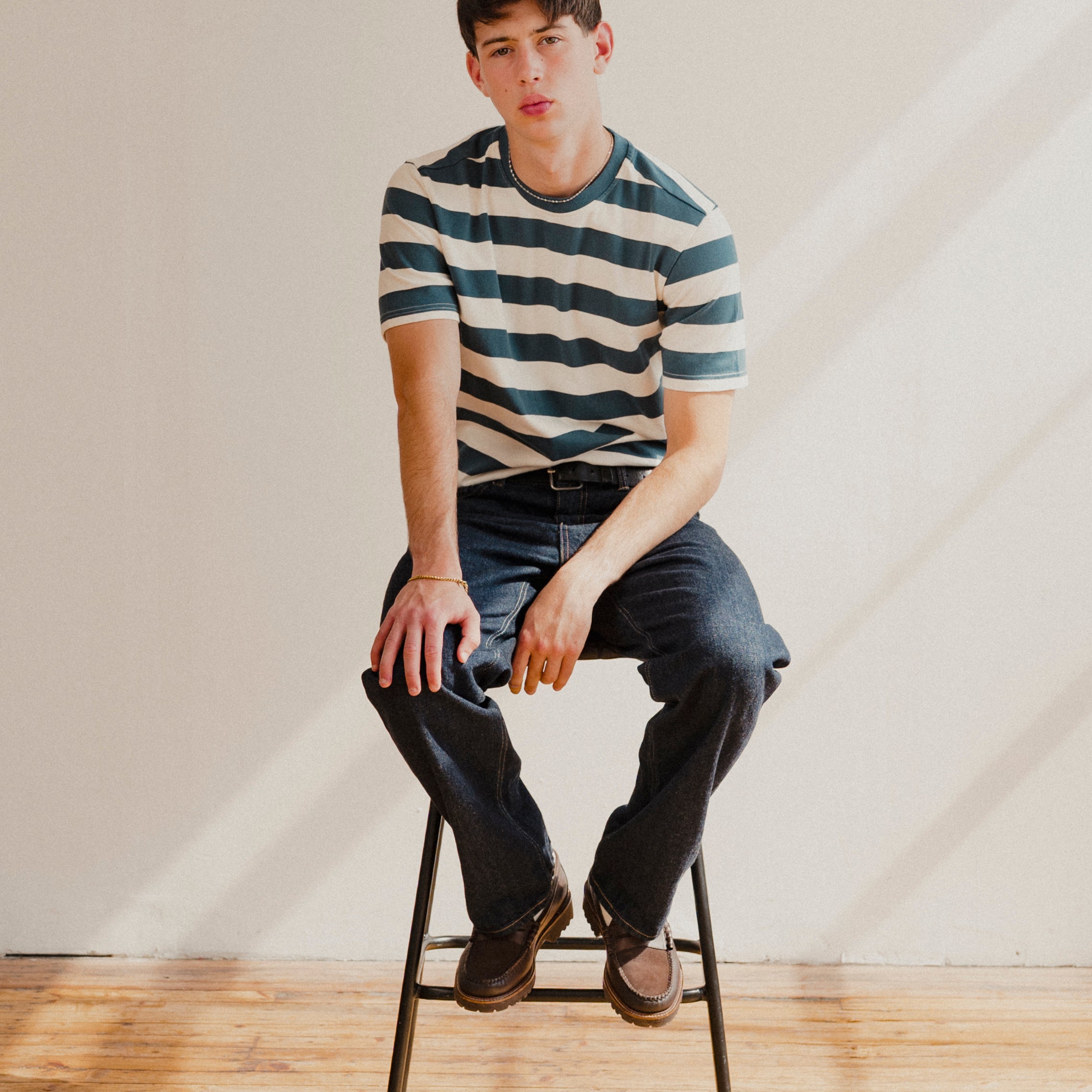 Man sitting on a stool wearing a blue and white striped t-shirt and jeans in a minimalistic setting.
