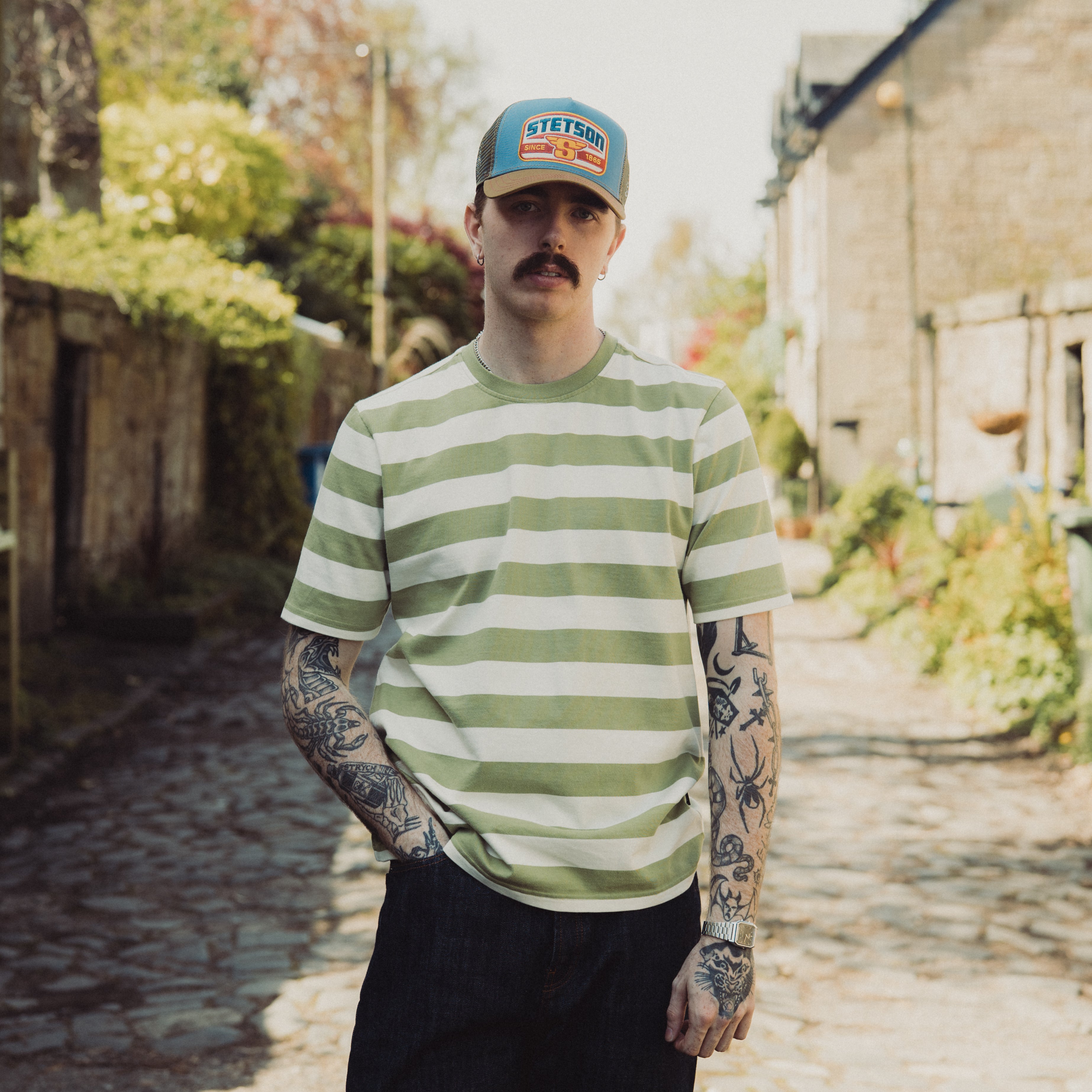 Man wearing a green and white striped shirt and cap standing in a sunlit alleyway.