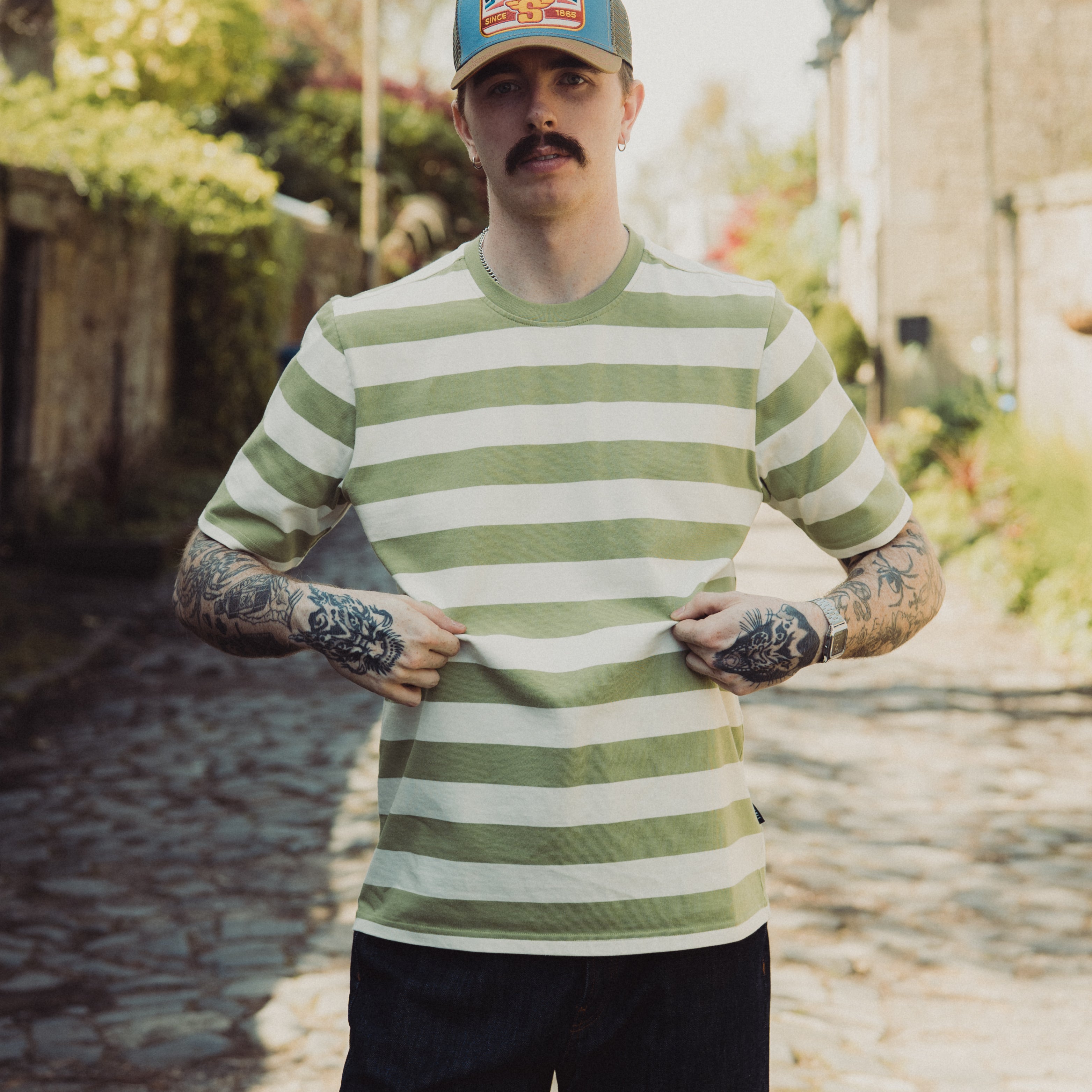 Man wearing a green and white striped shirt and a blue cap with 'Stetson' logo outdoors.