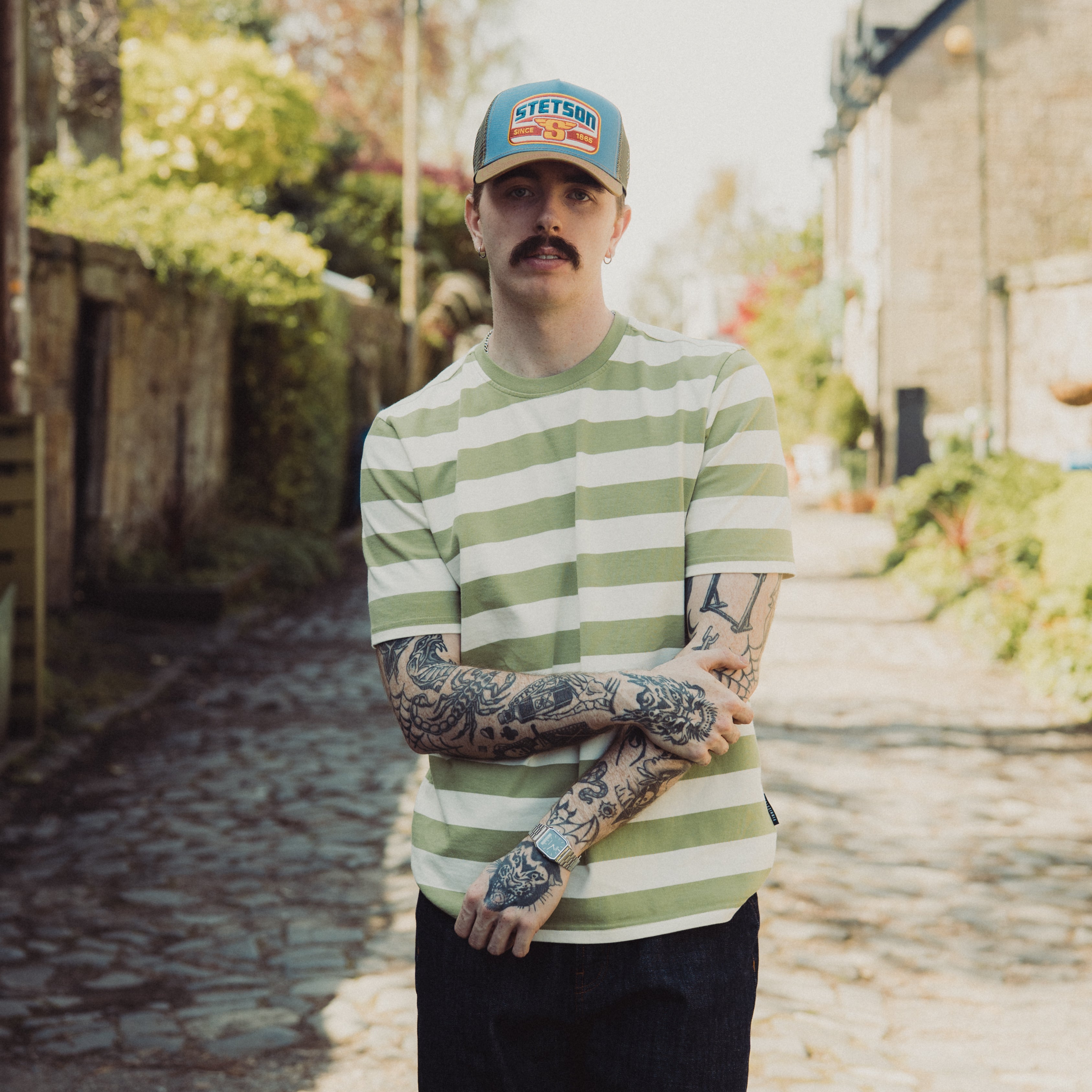 Man wearing a green and white striped shirt and cap with tattoos, standing outdoors.