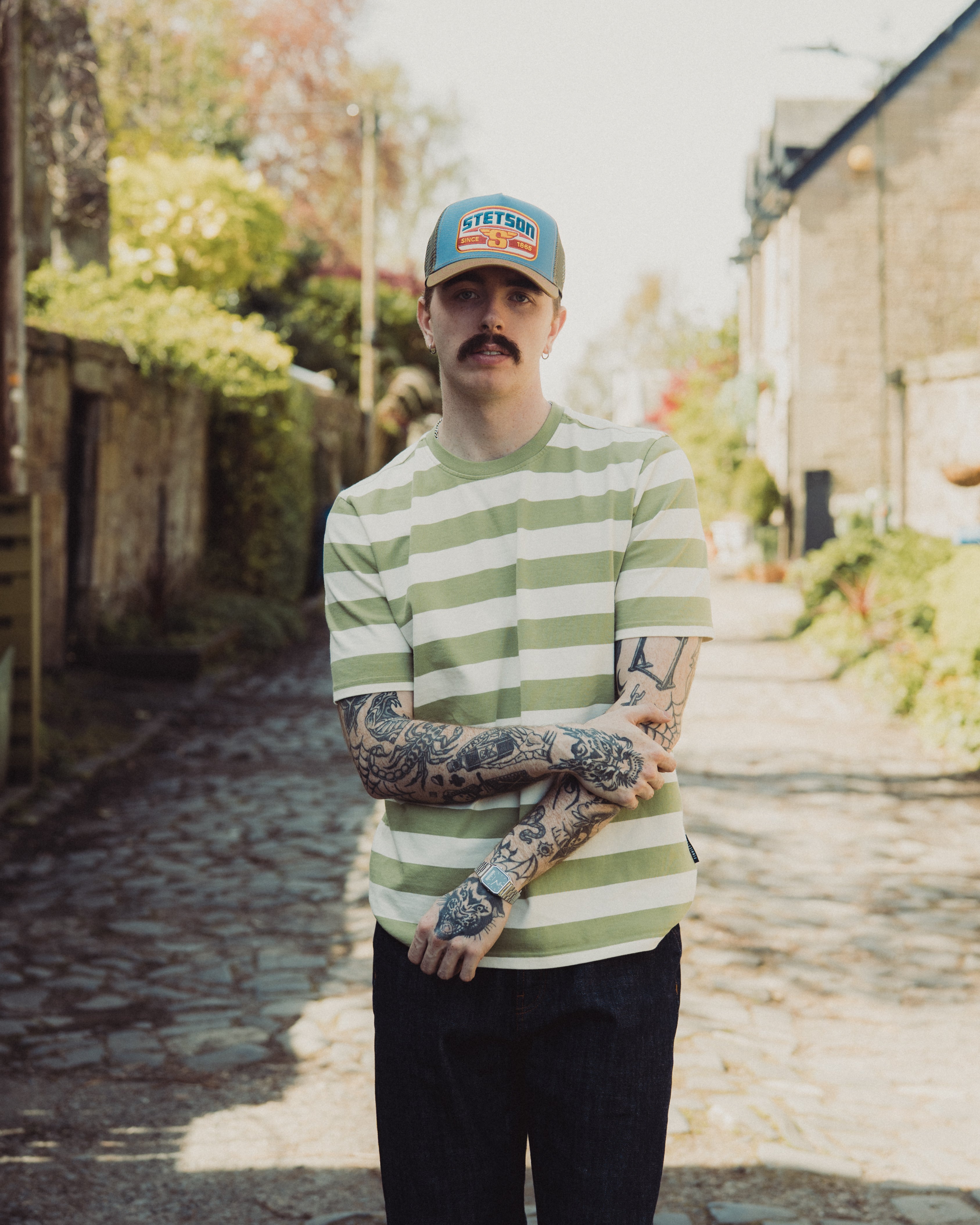 Man wearing a green and white striped shirt and cap with tattoos, standing outdoors.