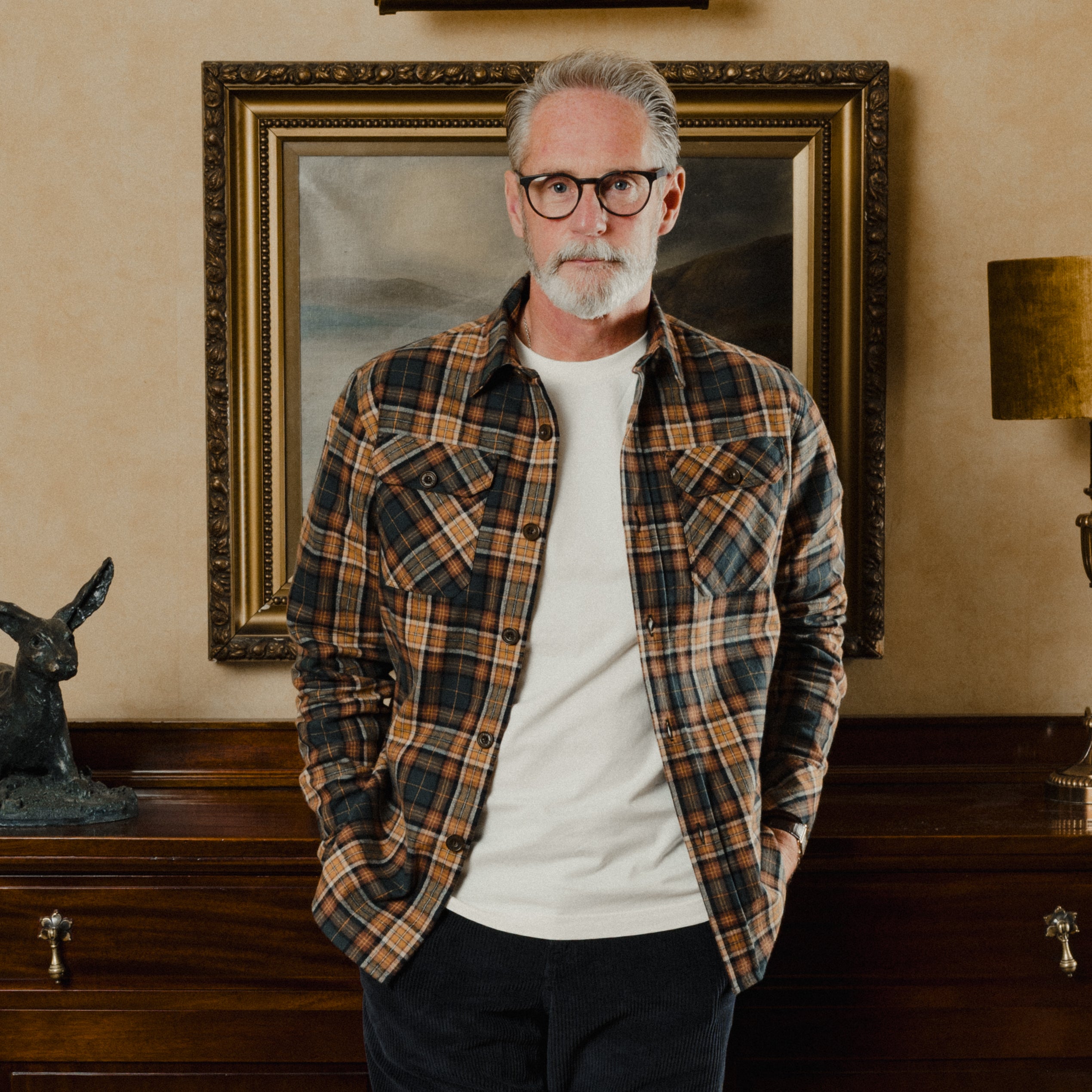 Man wearing a plaid shirt standing in a room with a painting on the wall.
