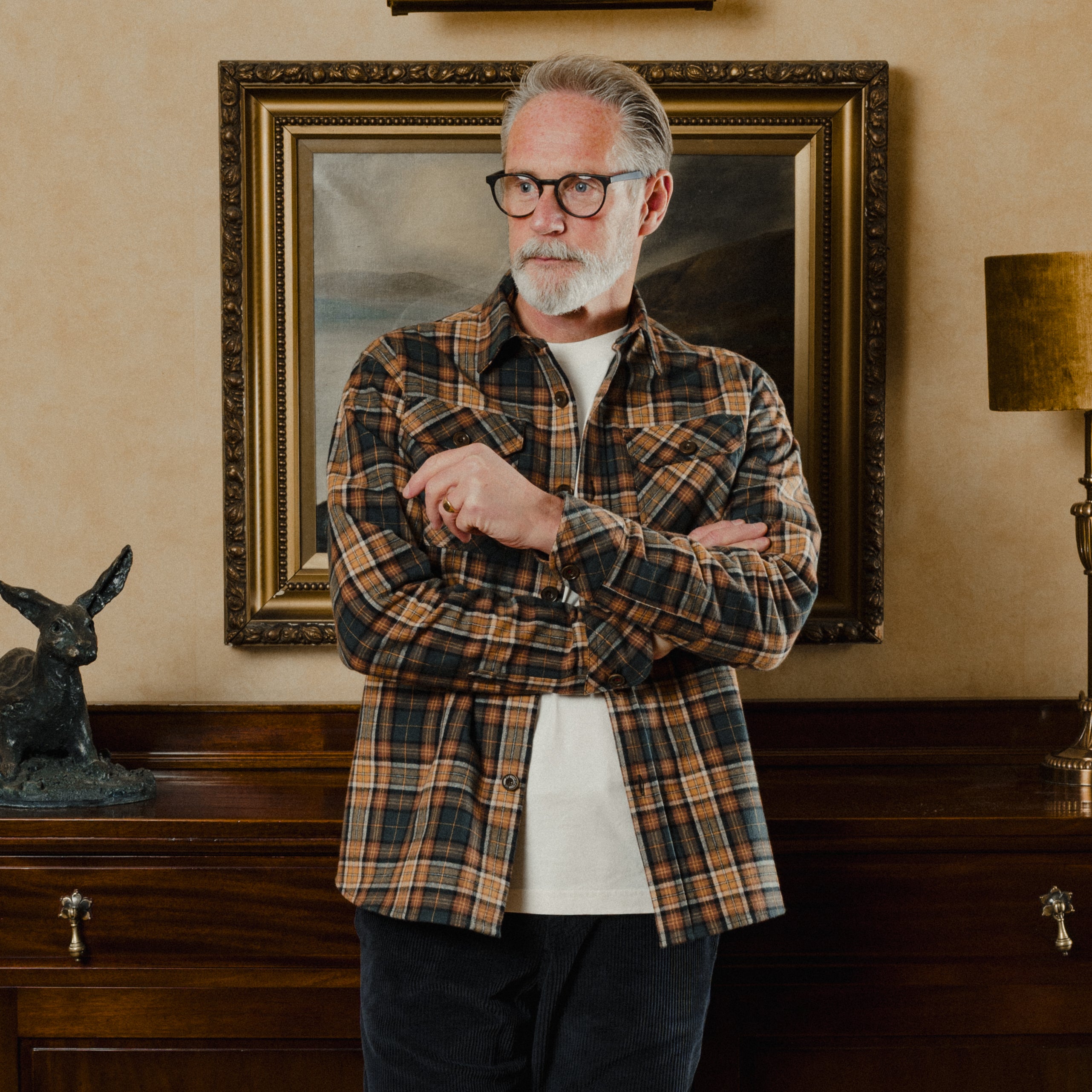 Man wearing a plaid shirt standing in a room with a painting and decorative items on the wall.