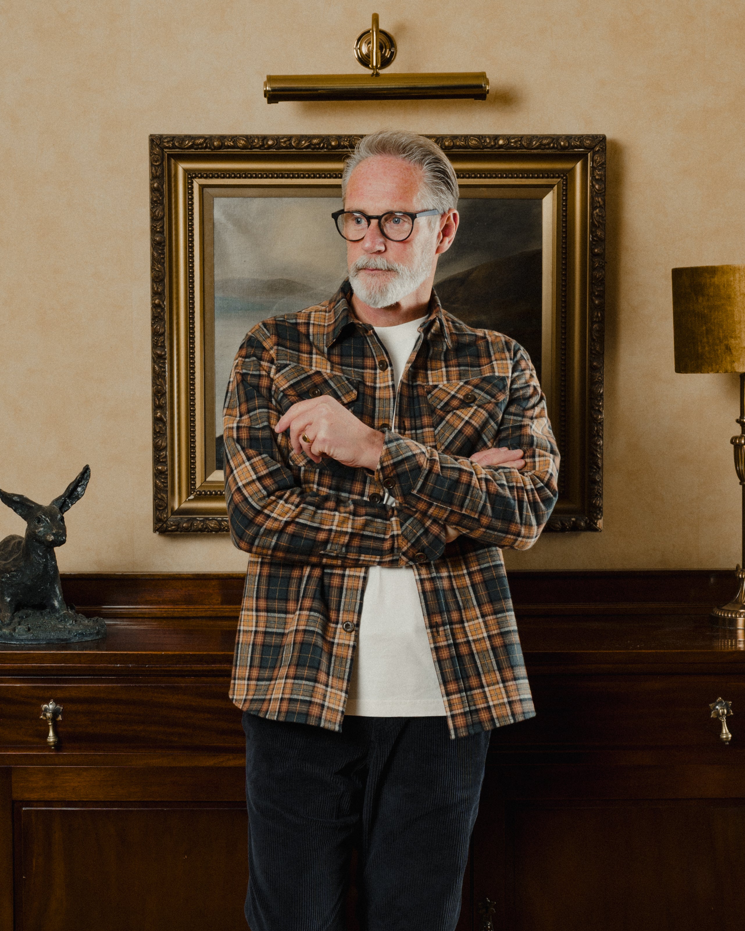 Man wearing a plaid shirt standing in a room with a painting and decorative items on the wall.
