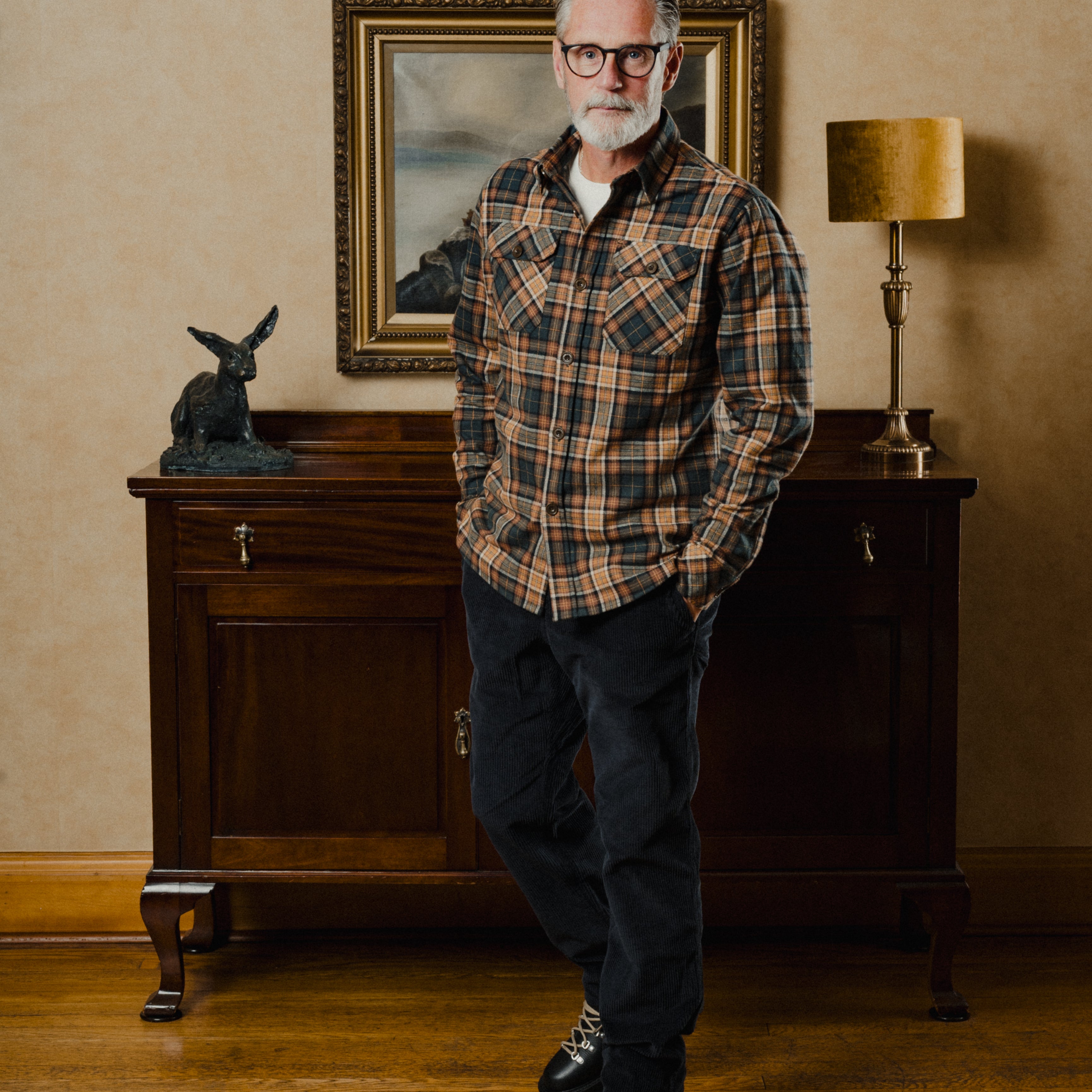 Man wearing a plaid shirt standing in a room with a wooden cabinet and lamp.
