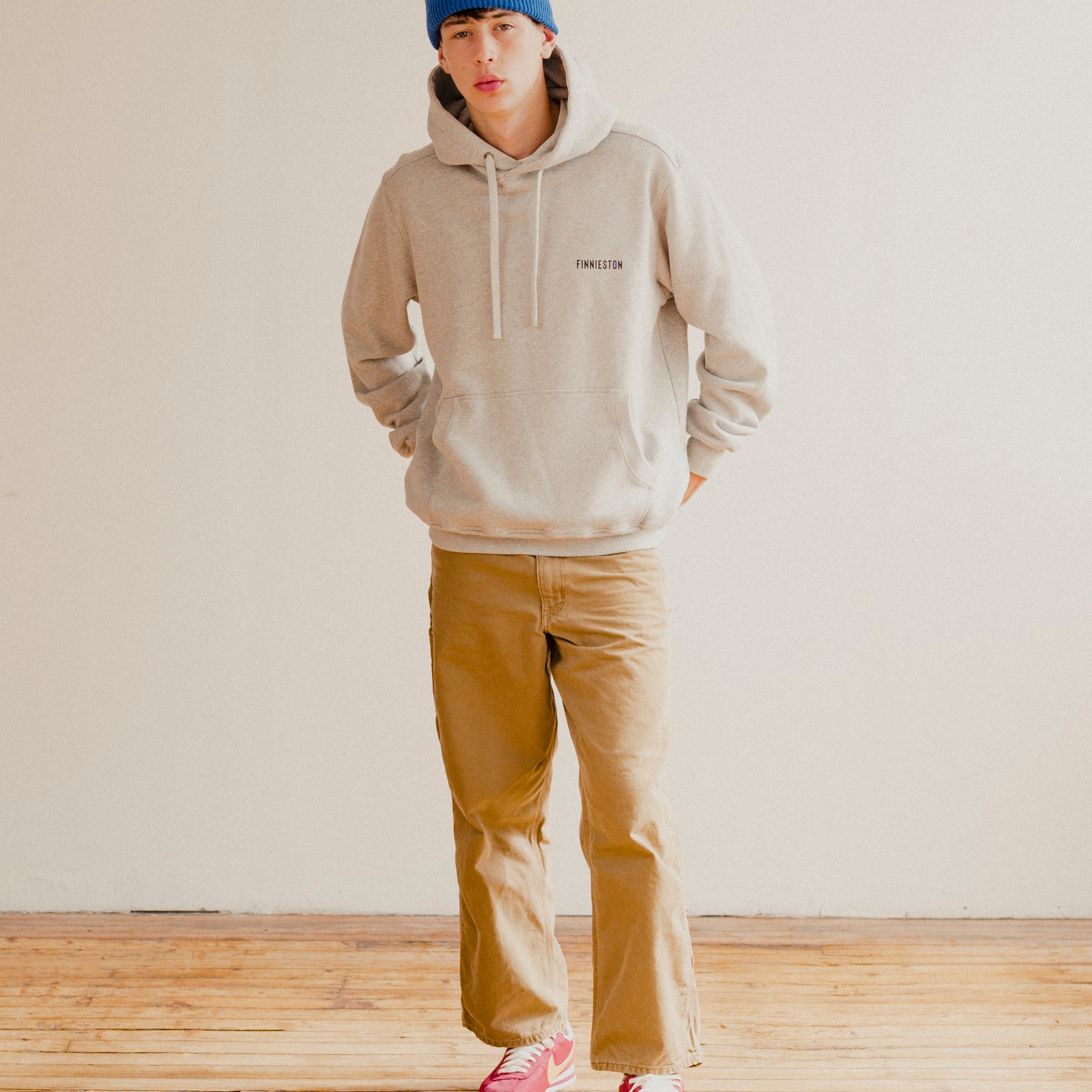 Person wearing a grey hoodie, blue beanie, and tan pants standing in a room with wooden flooring.