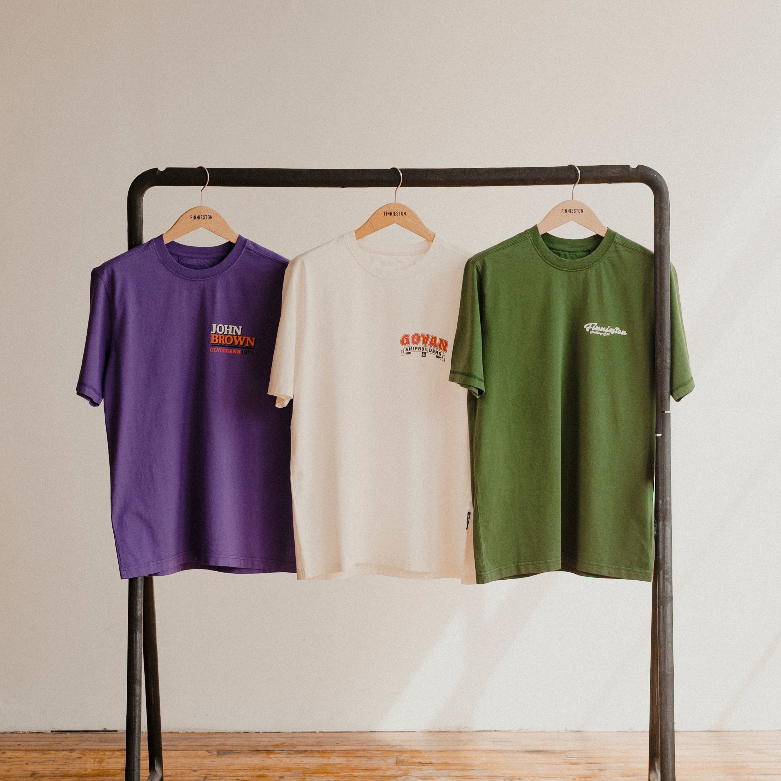 Three t-shirts in purple, white, and green hanging on a rack against a plain background.