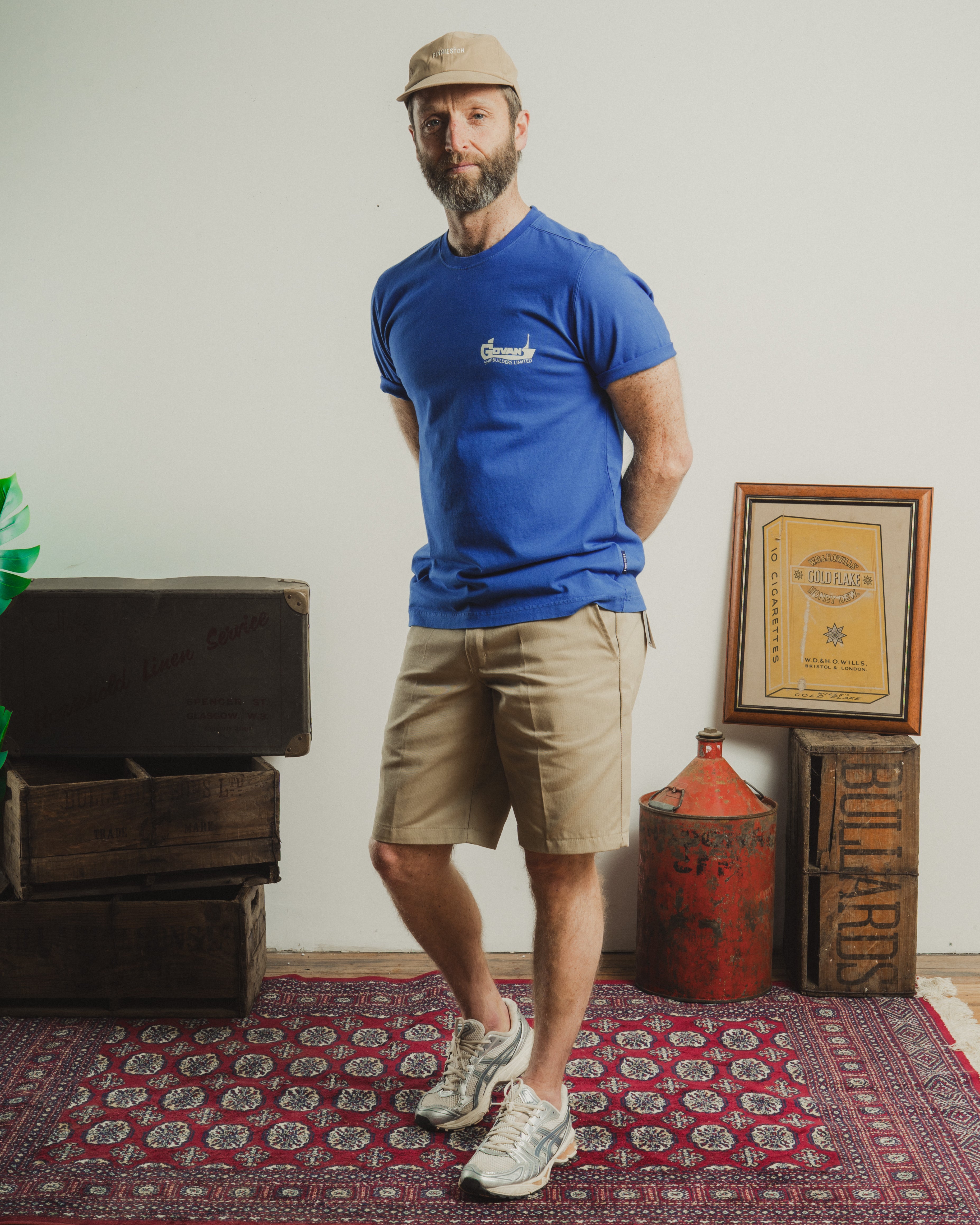 Man wearing a blue t-shirt with a logo, beige shorts, and a cap standing in a room with wooden furniture and a framed document on the wall.