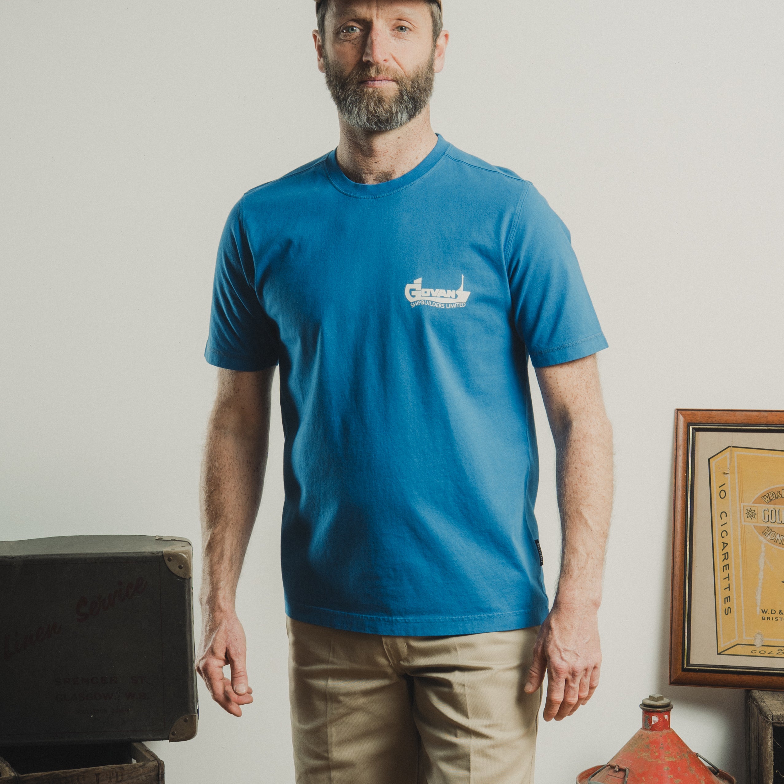Man wearing a blue t-shirt with a logo, beige cap, and shorts standing in a rustic setting.
