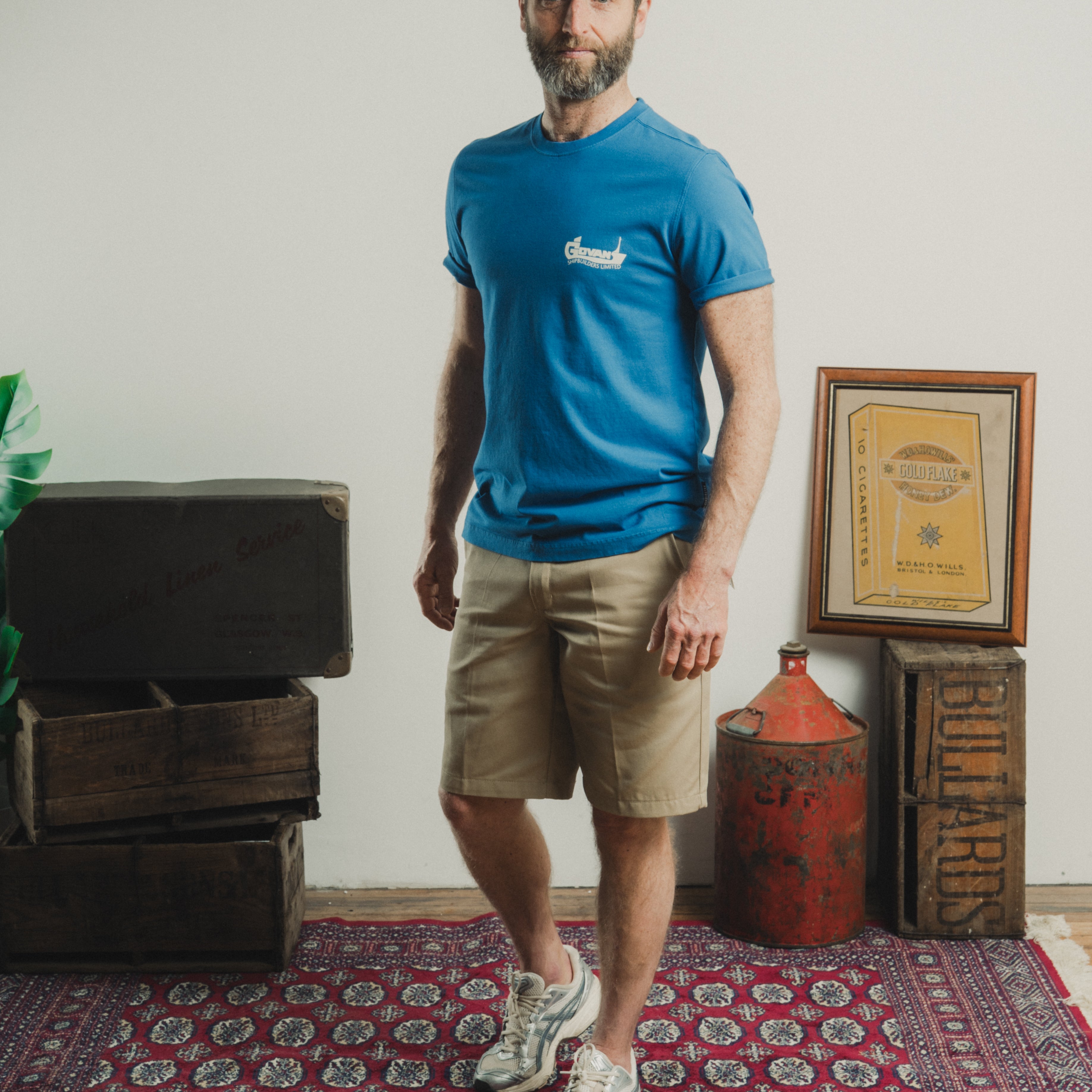 Man wearing a blue t-shirt and beige shorts standing in a room with a rug and wooden crates.