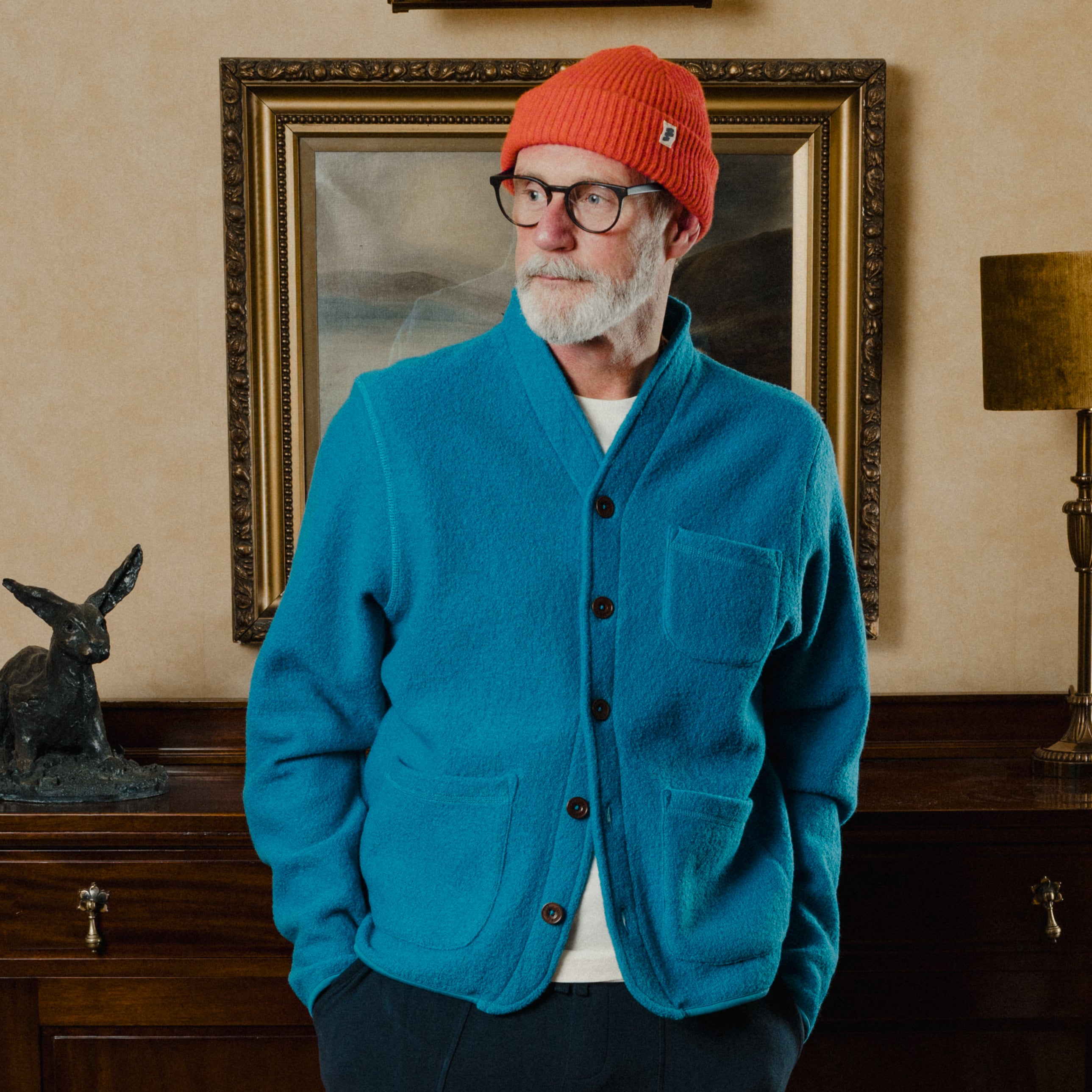 Man wearing a blue cardigan and red beanie standing in a room with a painting and lamp.