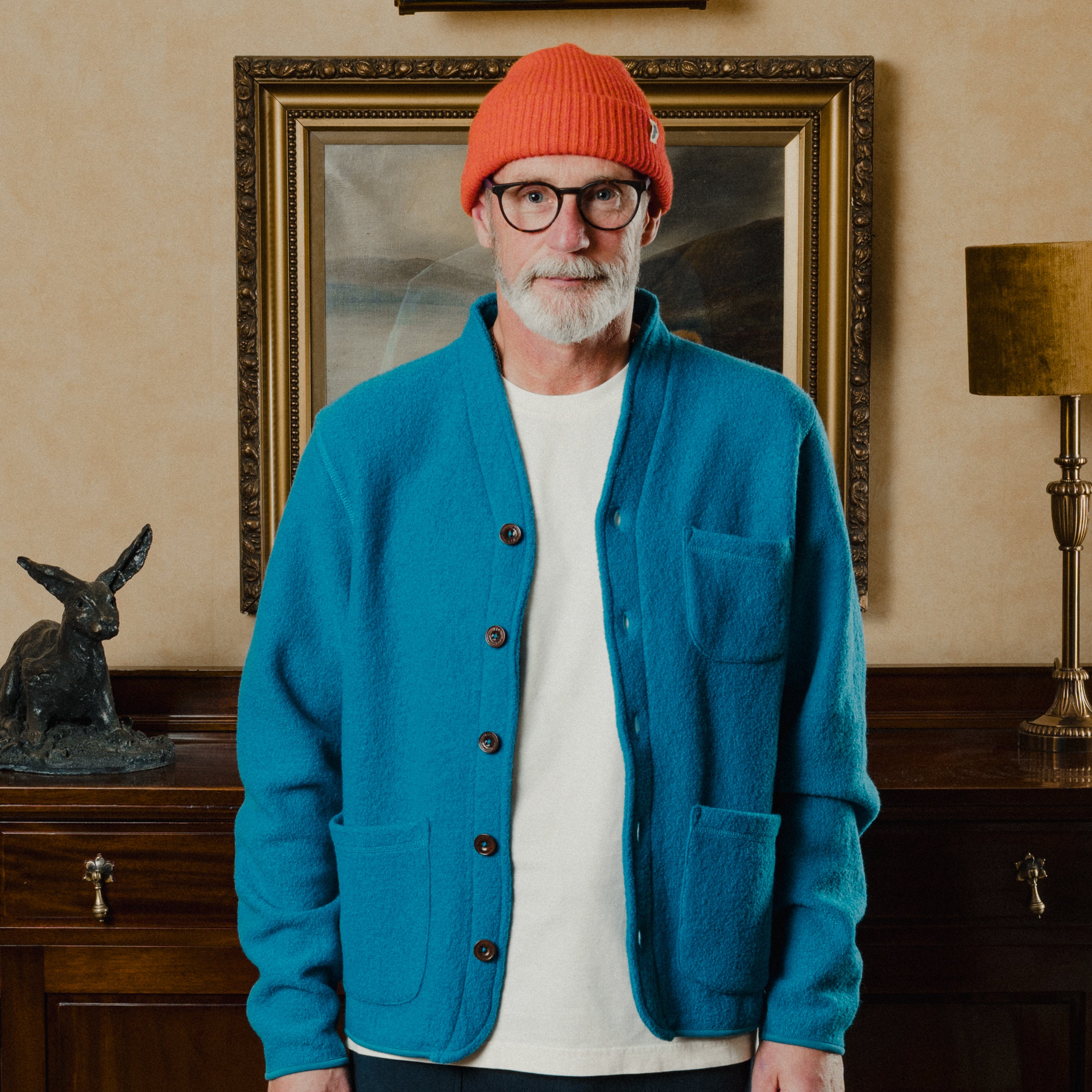 Man wearing a blue cardigan, white shirt, and orange beanie standing in a room with a painting and lamp.