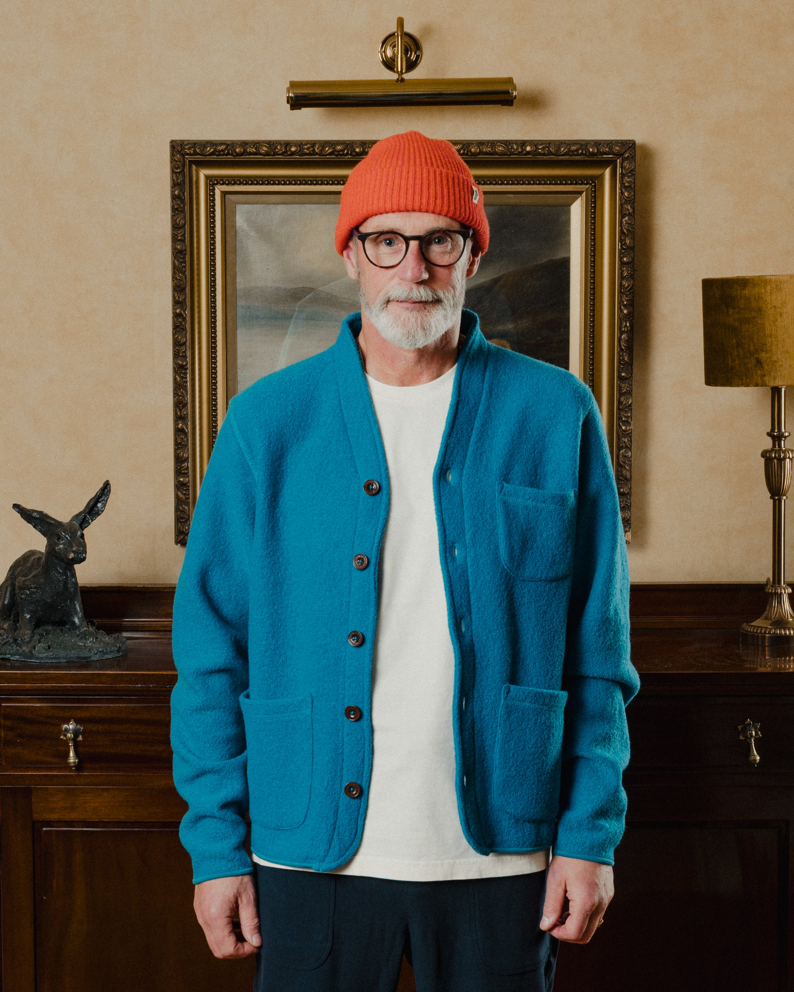 Man wearing a blue cardigan, white shirt, and orange beanie standing in a room with a painting and lamp.