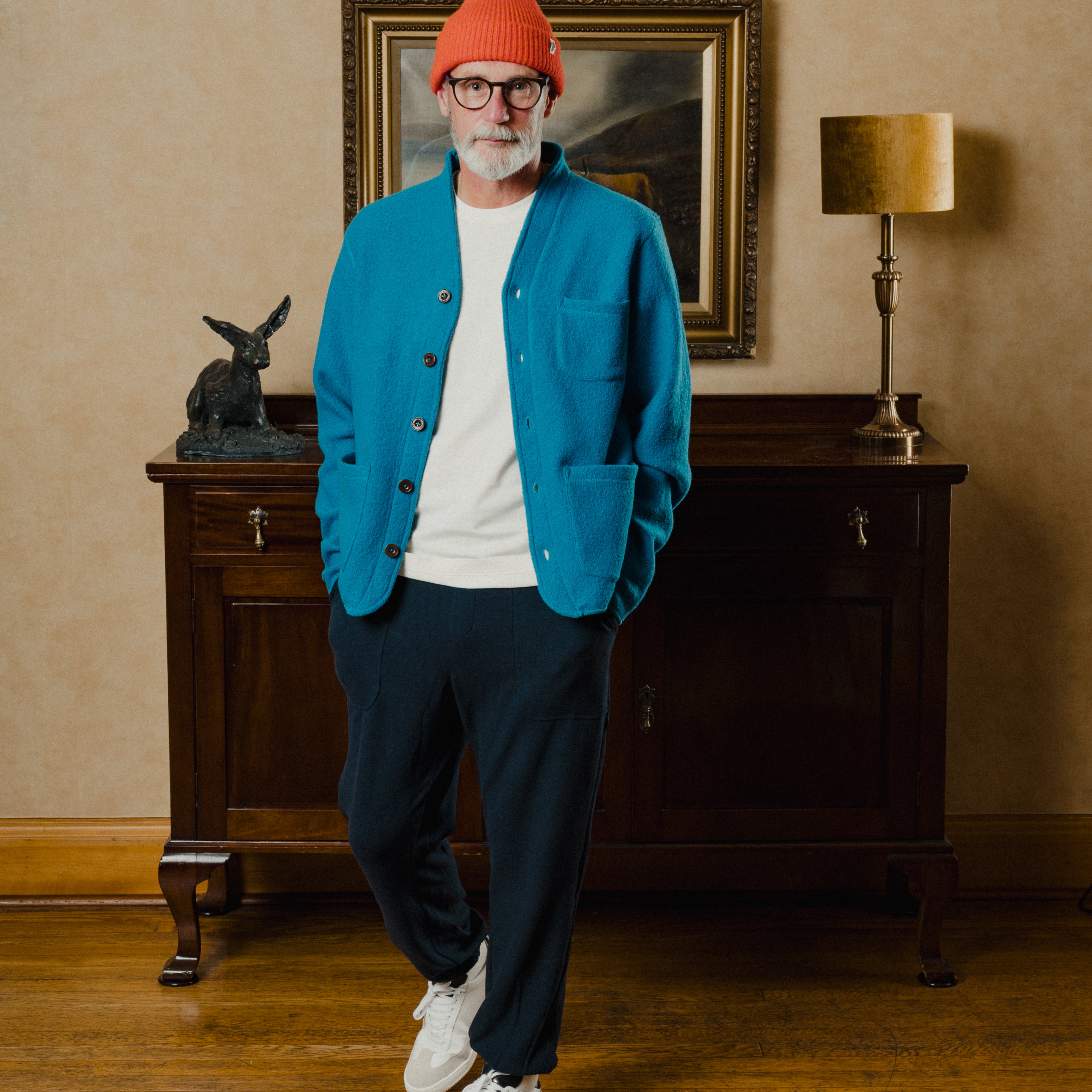 Man wearing a blue jacket, white shirt, navy pants, and orange beanie standing in a room with a lamp and painting.