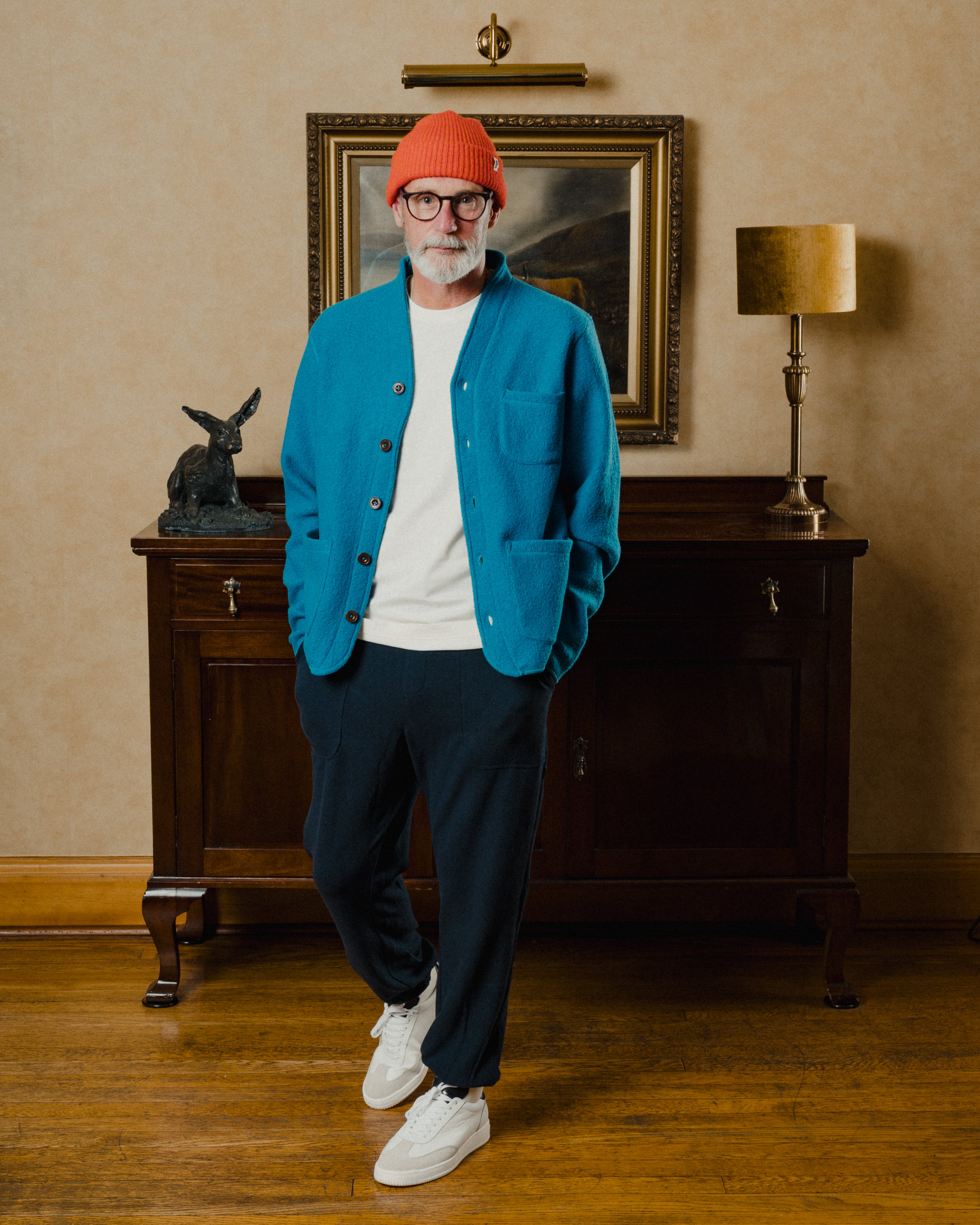Man wearing a blue jacket, white shirt, navy pants, and orange beanie standing in a room with a lamp and painting.