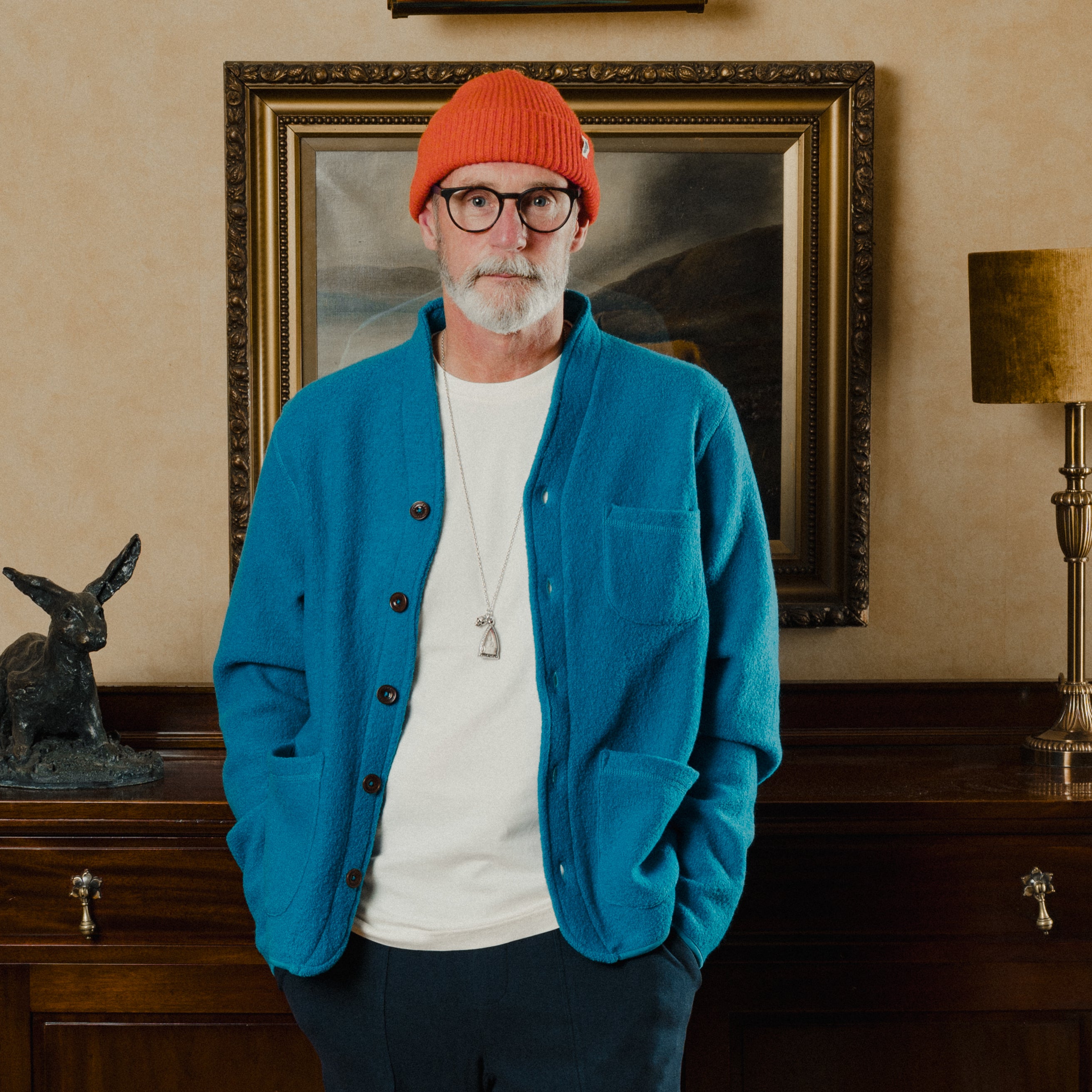 Man wearing a blue jacket and red beanie standing in a room with a painting and lamp.