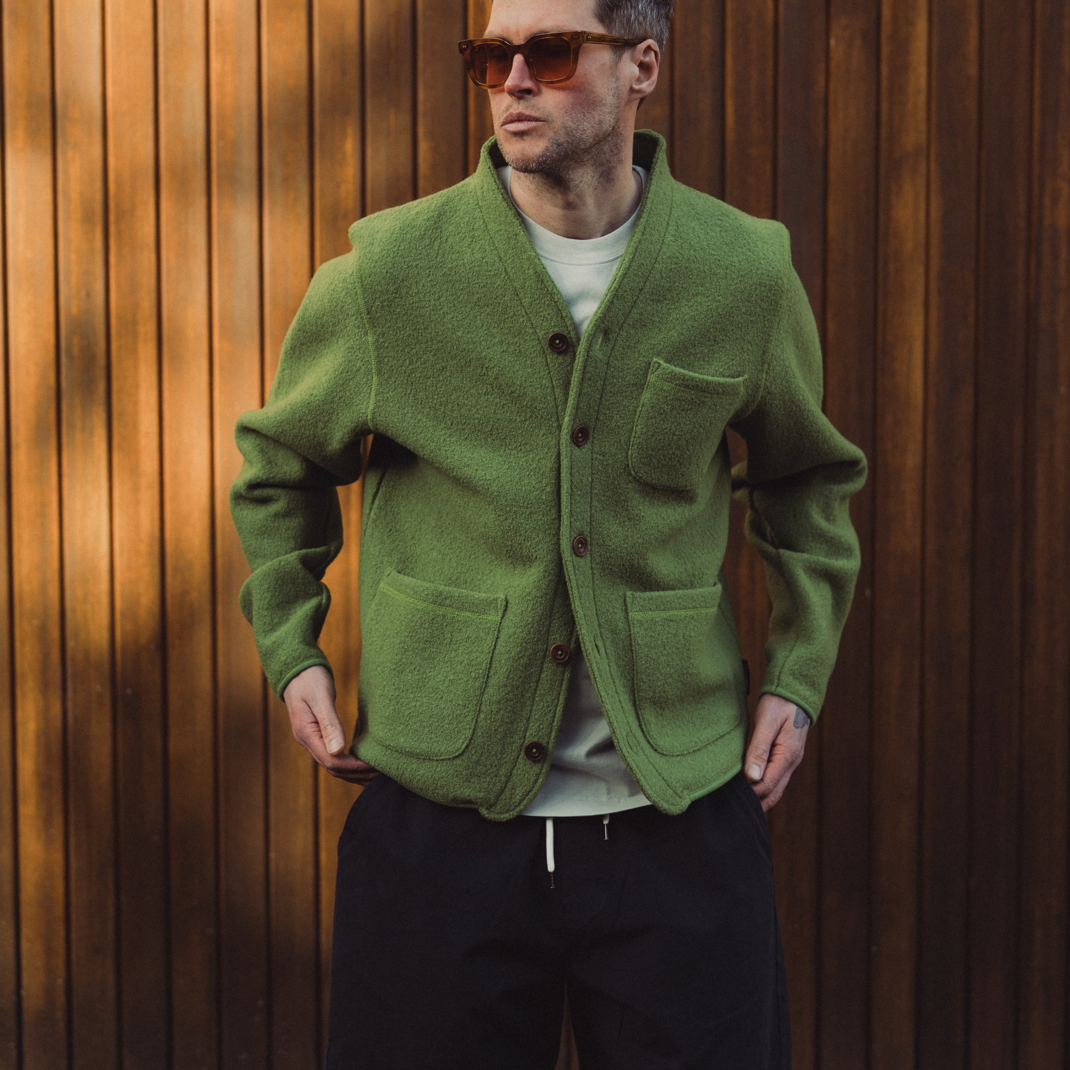 Man wearing a green cardigan, sunglasses, and black shorts against a wooden background