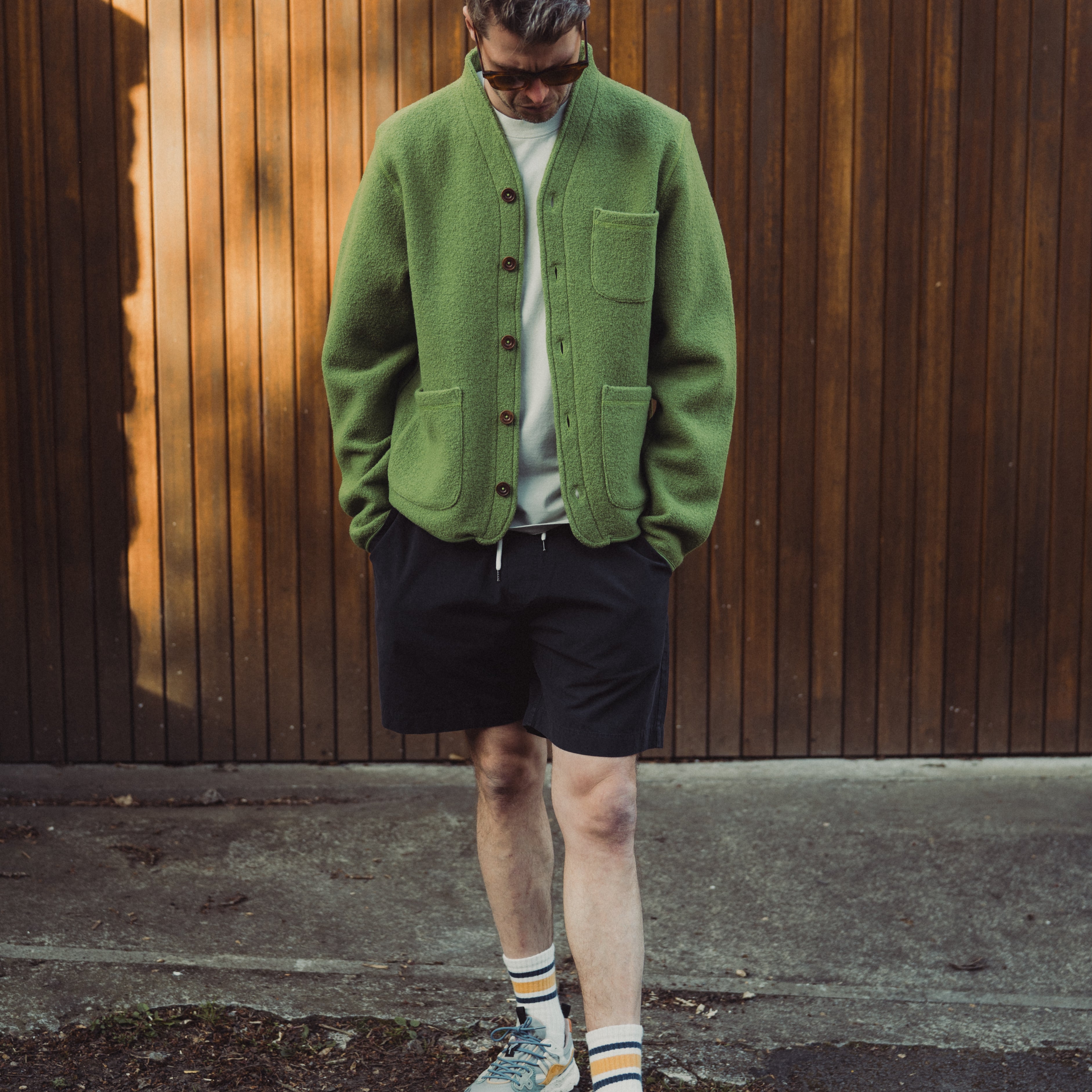 Man wearing a green cardigan, black shorts, and colorful sneakers against a wooden wall.
