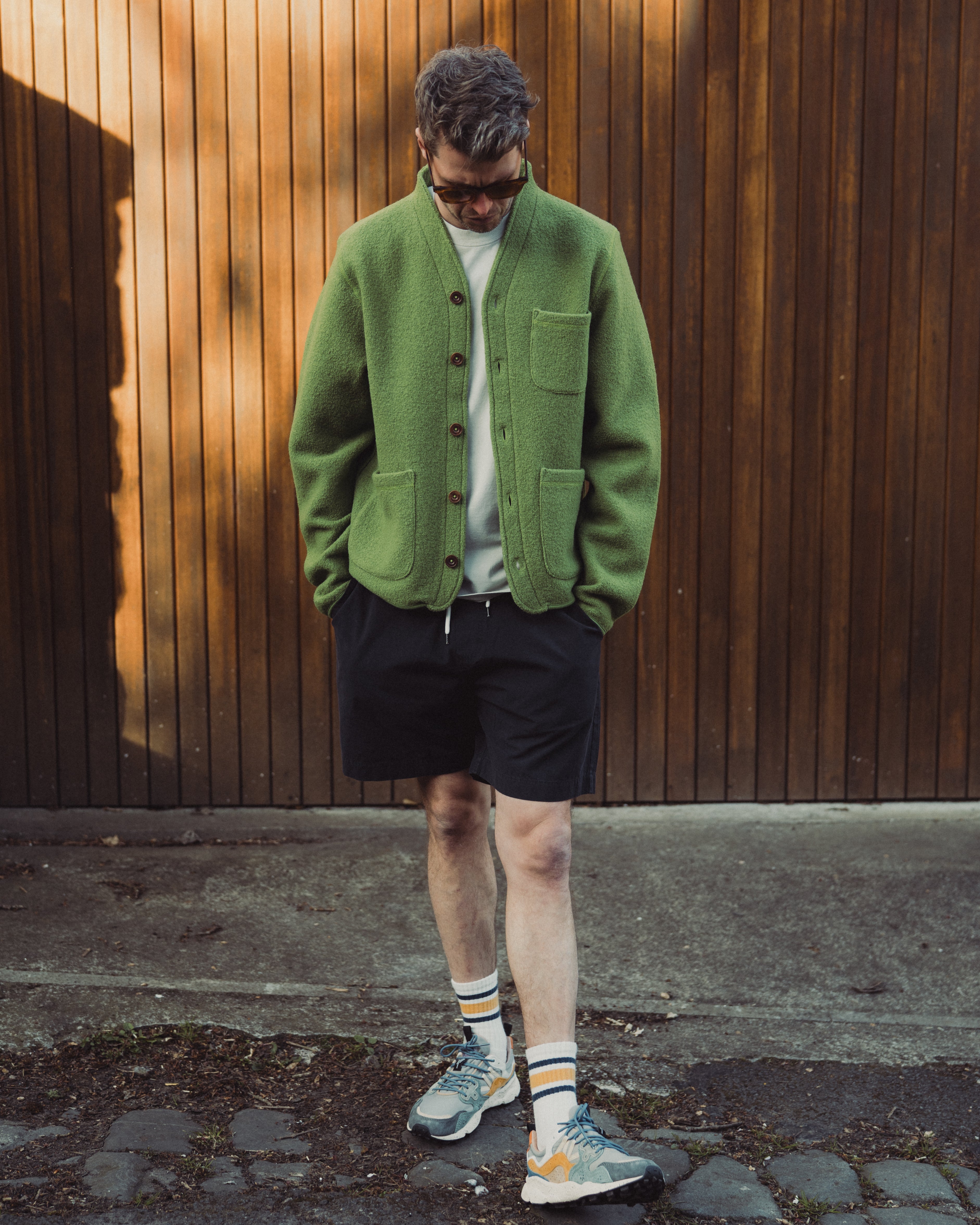 Man wearing a green cardigan, black shorts, and colorful sneakers against a wooden wall.