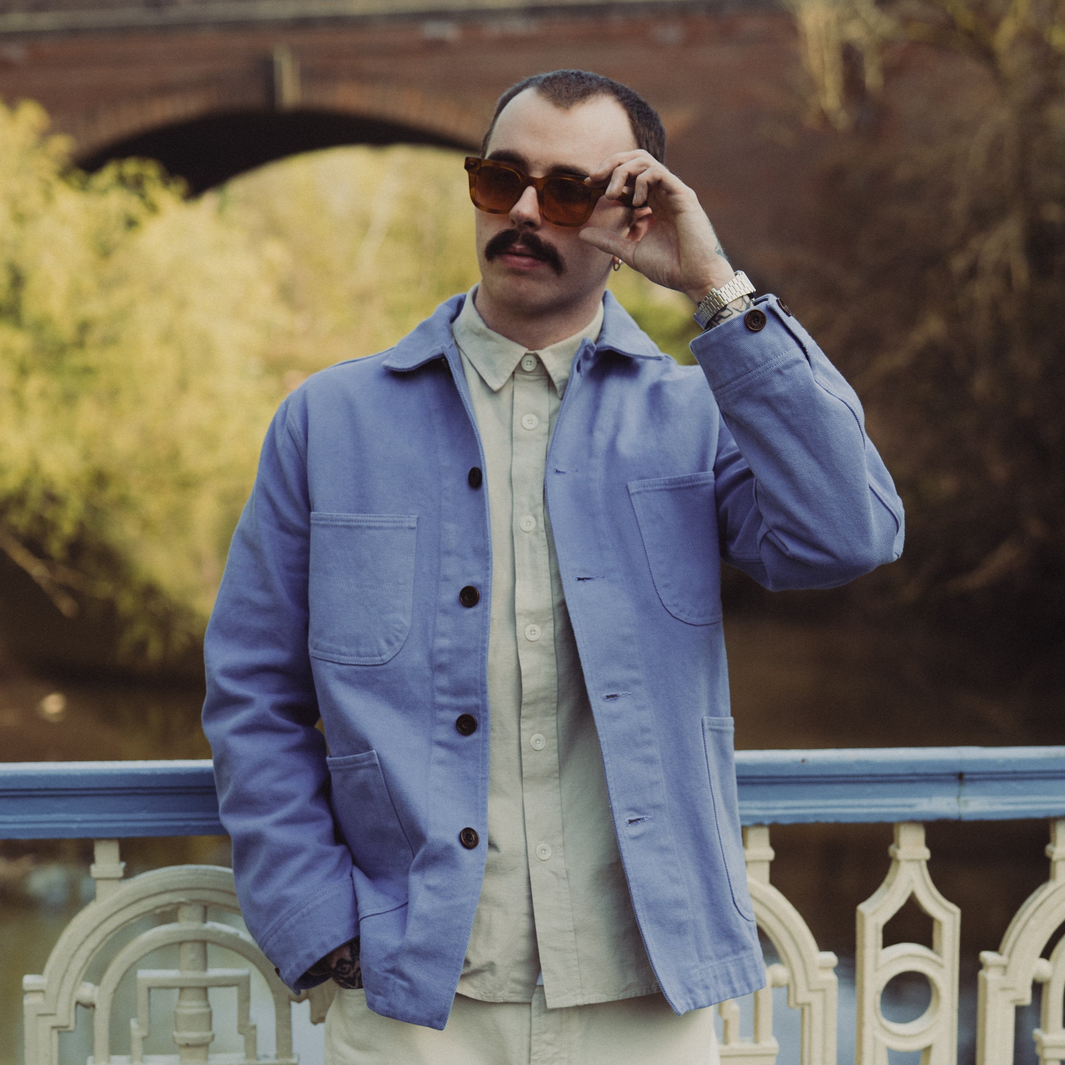 Man wearing a blue jacket and sunglasses, standing by a railing with a bridge in the background.