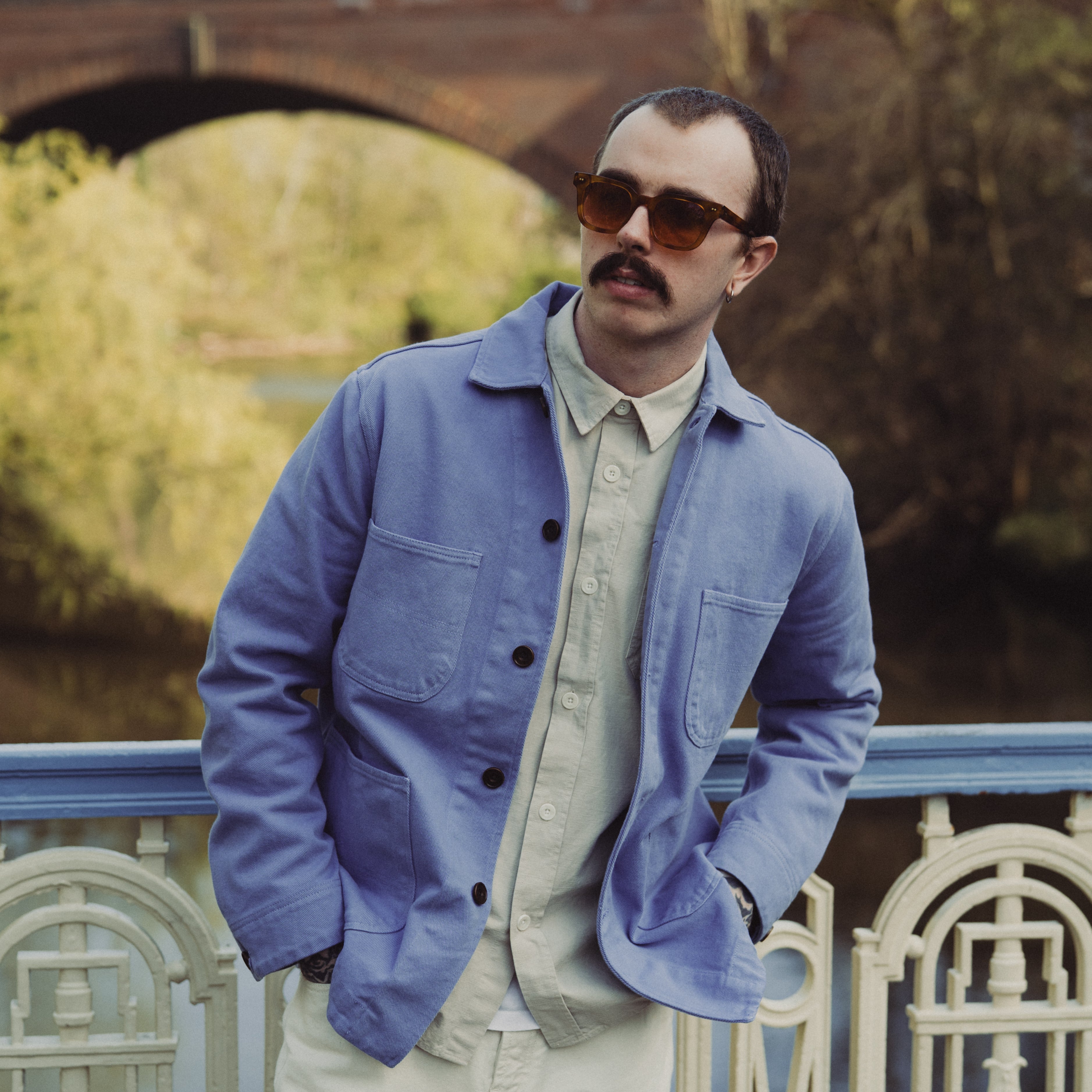 Man wearing a blue jacket and sunglasses standing by a railing with a bridge and trees in the background