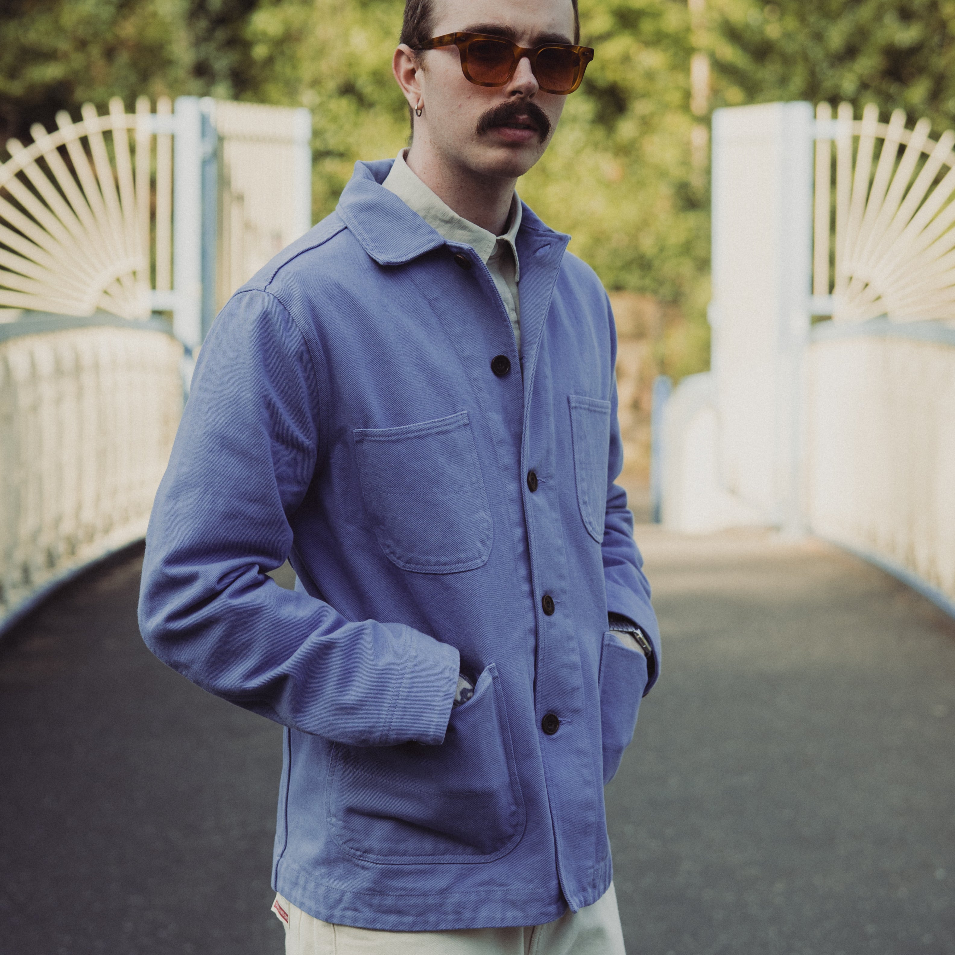 Man wearing a blue jacket and sunglasses standing on a path with greenery in the background