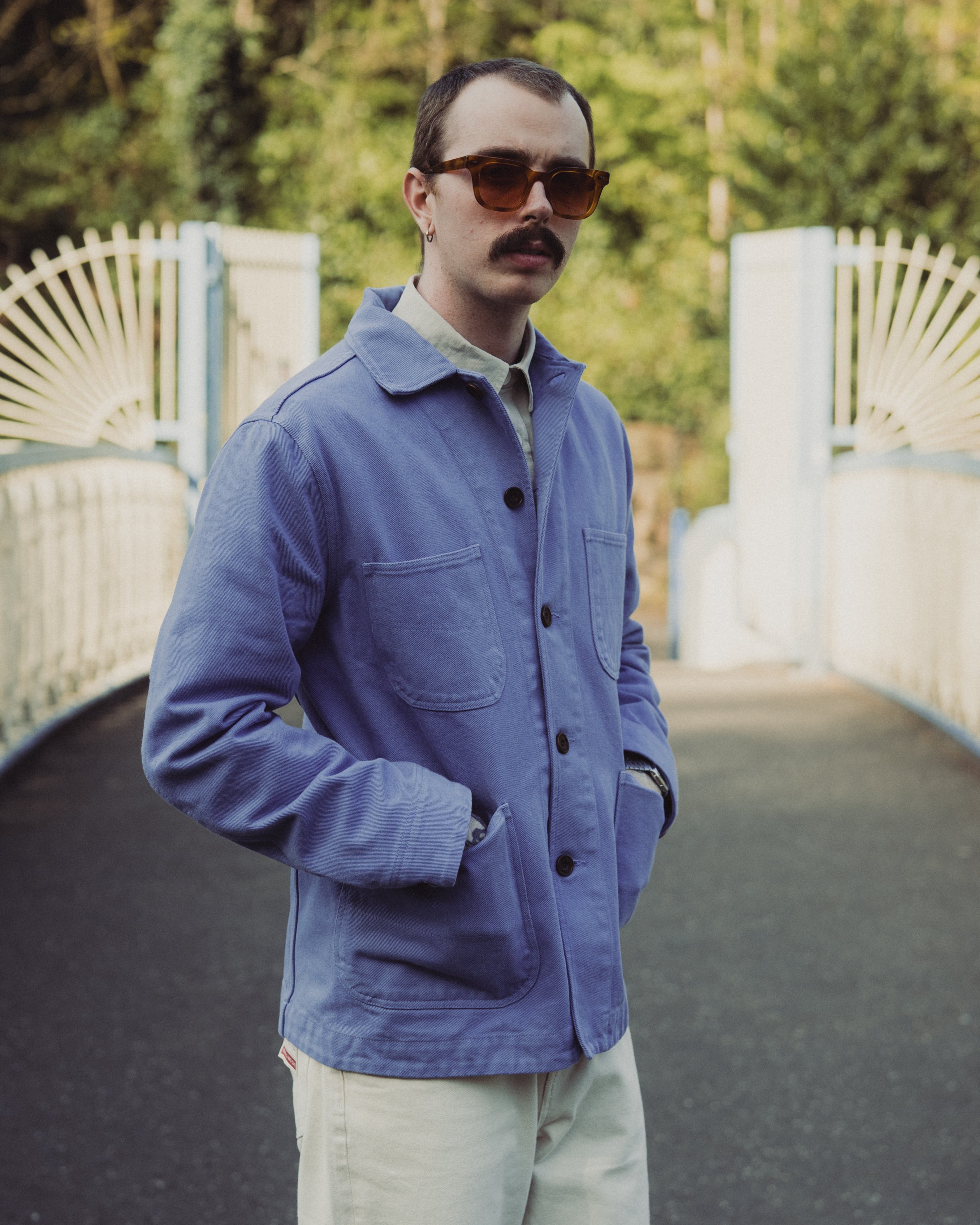 Man wearing a blue jacket and sunglasses standing on a path with greenery in the background