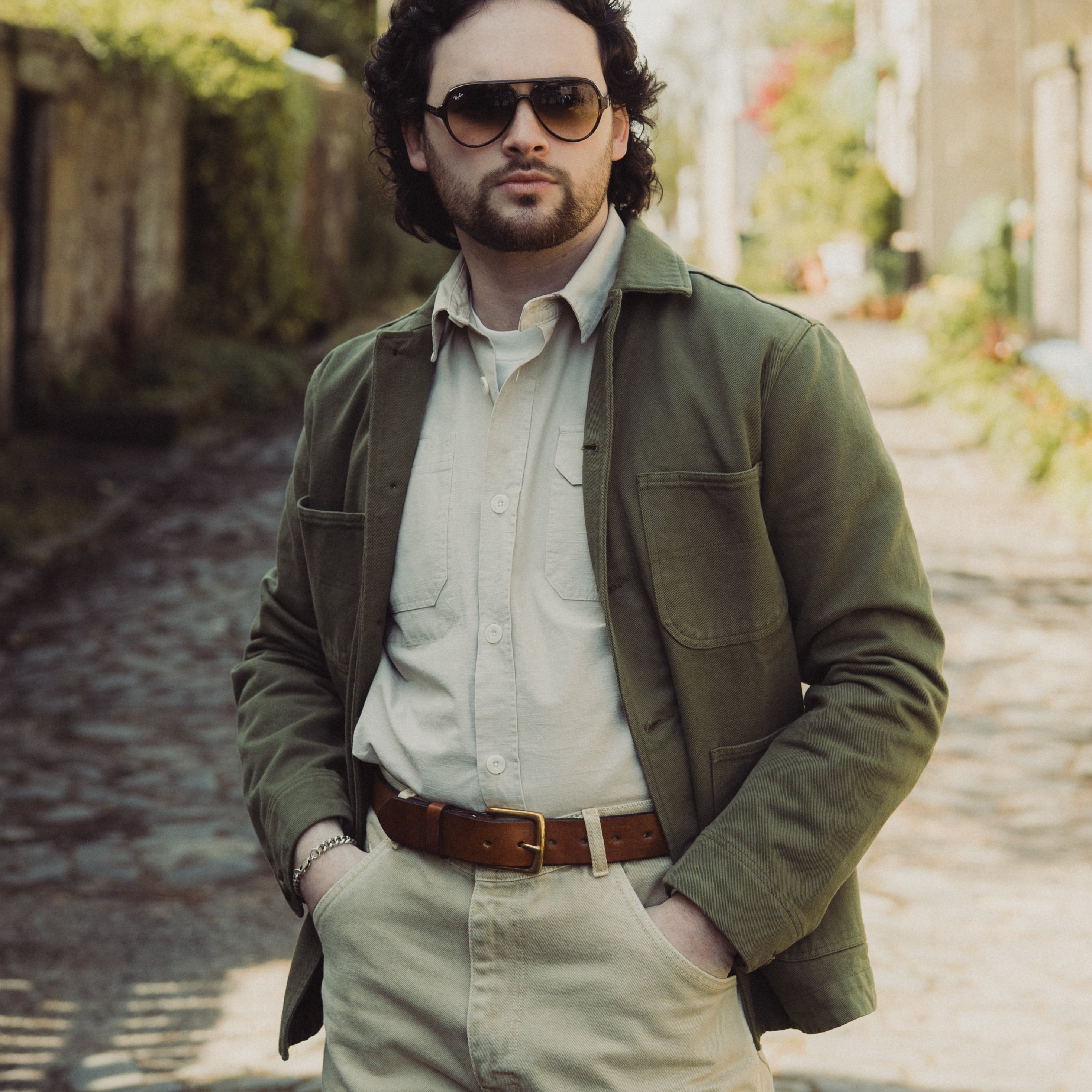 Man wearing sunglasses and a green jacket standing on a stone path with blurred background
