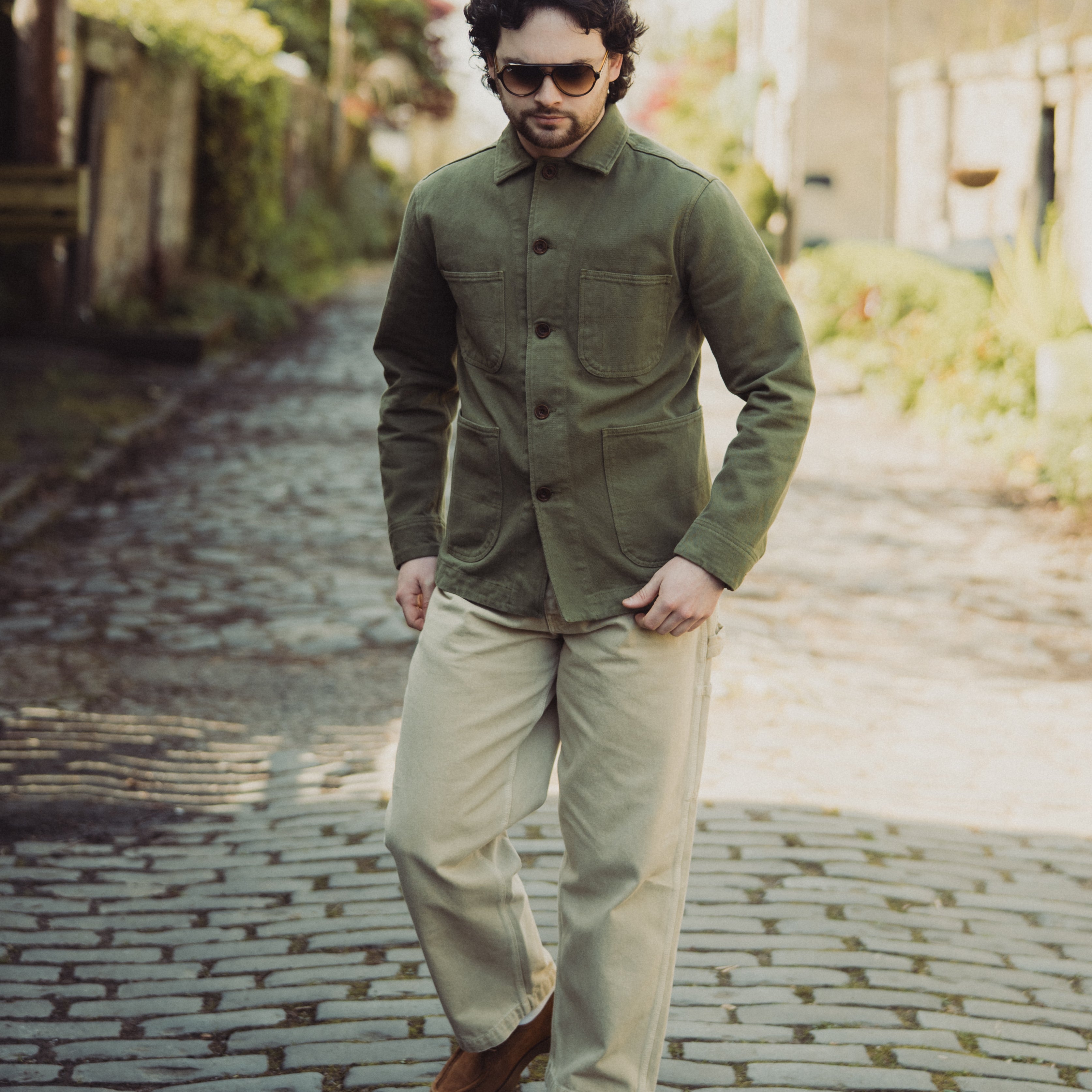 Man wearing a green jacket and beige pants walking on a cobblestone street.