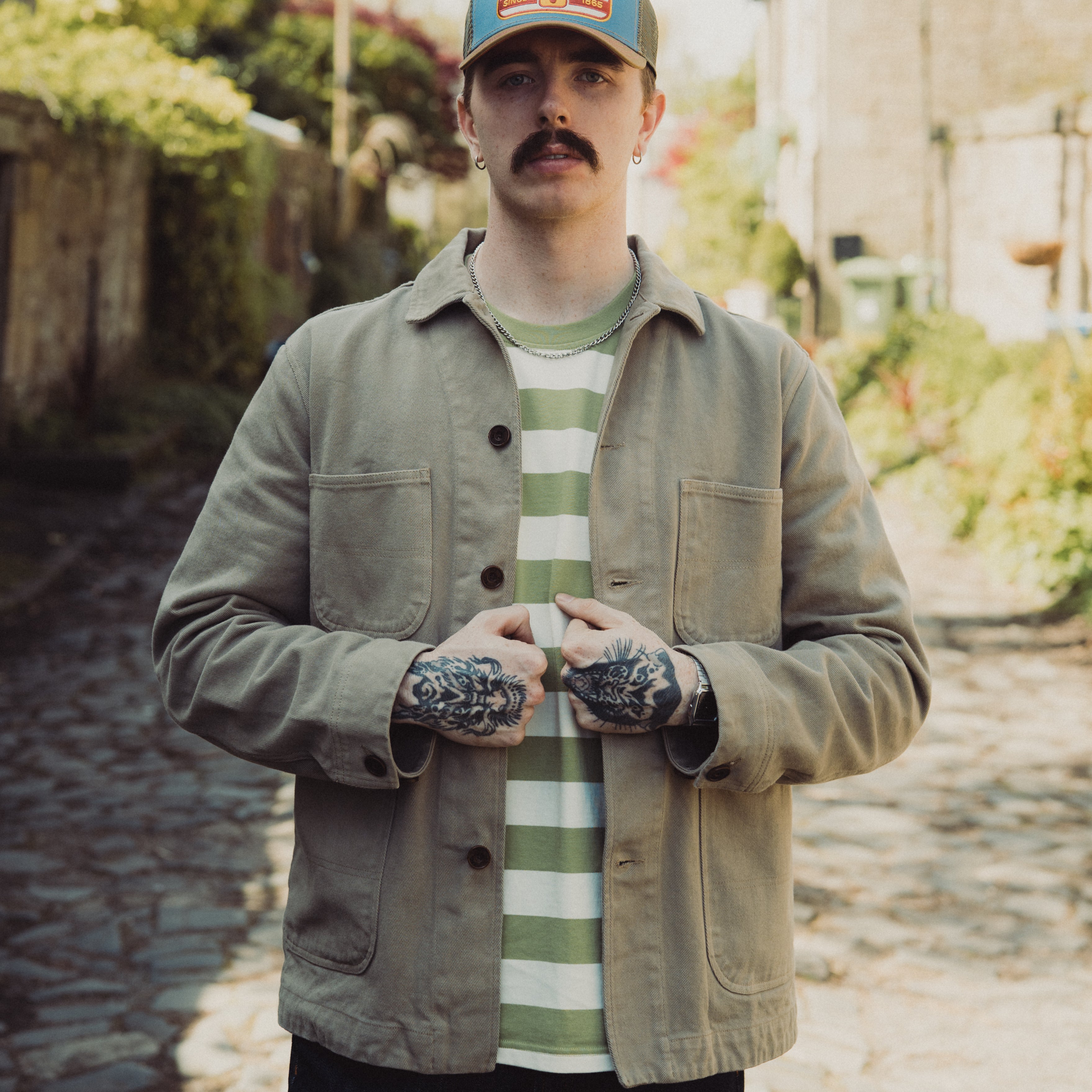 Man wearing a cap and jacket standing on a stone path with greenery