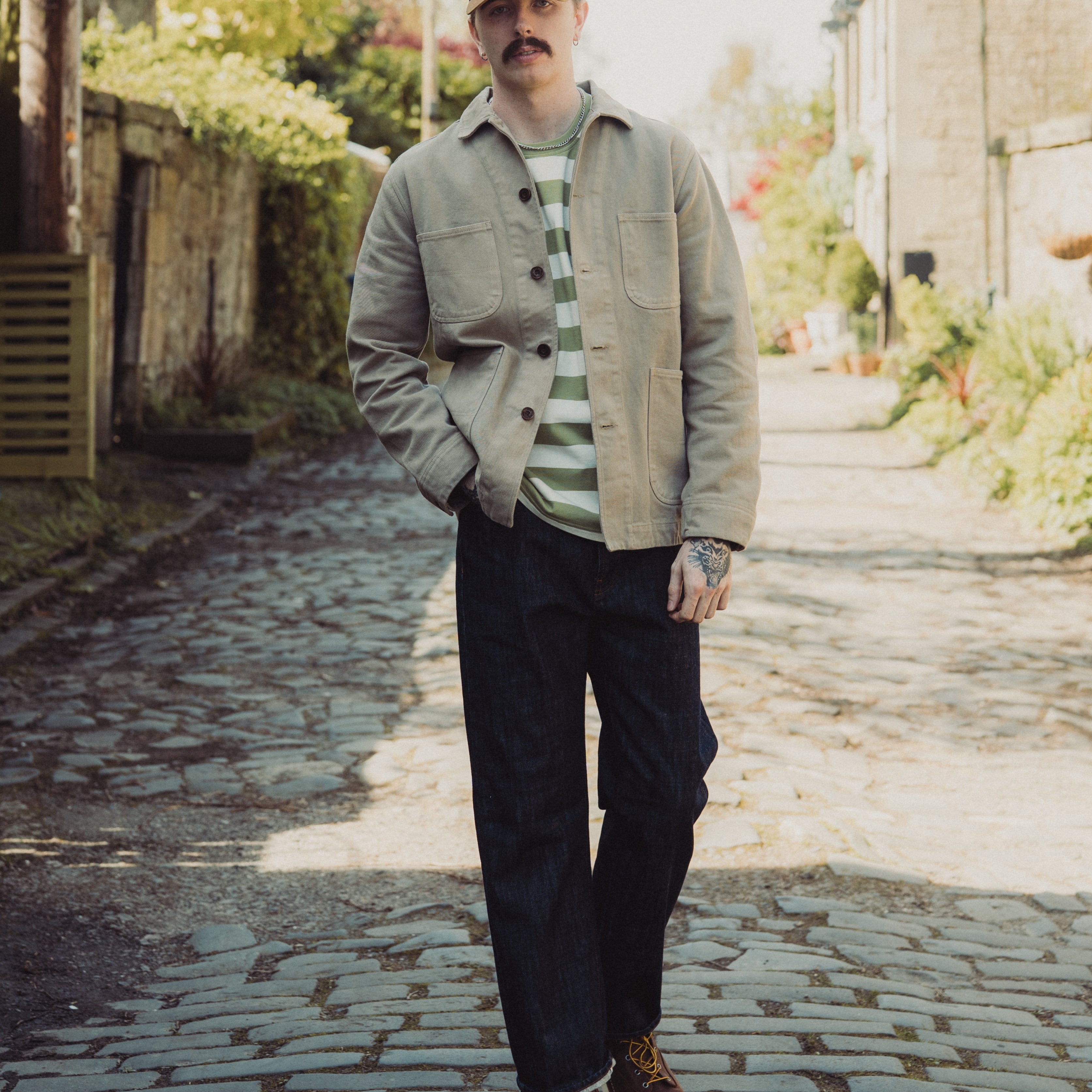 Man standing on a cobblestone path wearing a cap, jacket, and striped shirt.