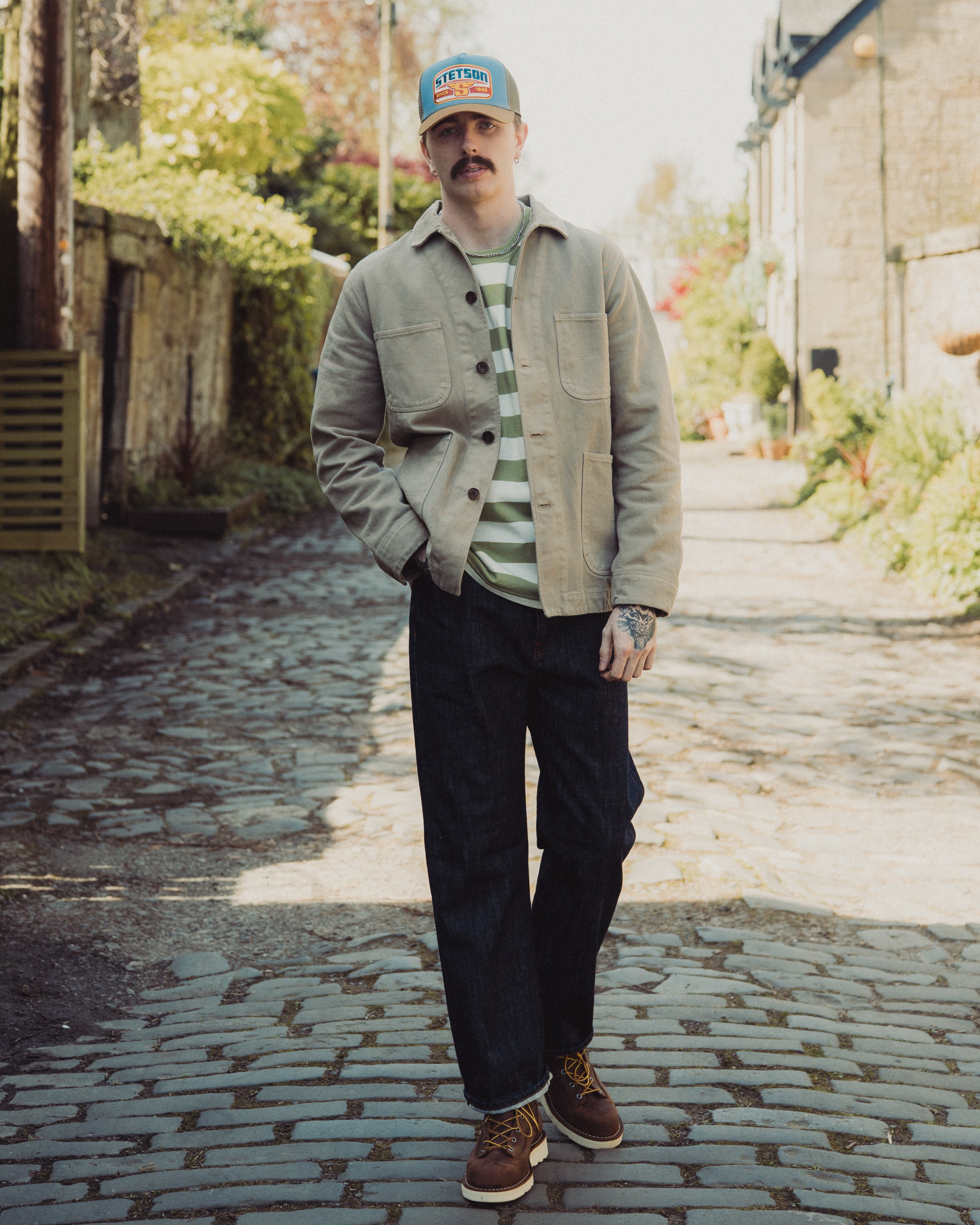 Man standing on a cobblestone path wearing a cap, jacket, and striped shirt.