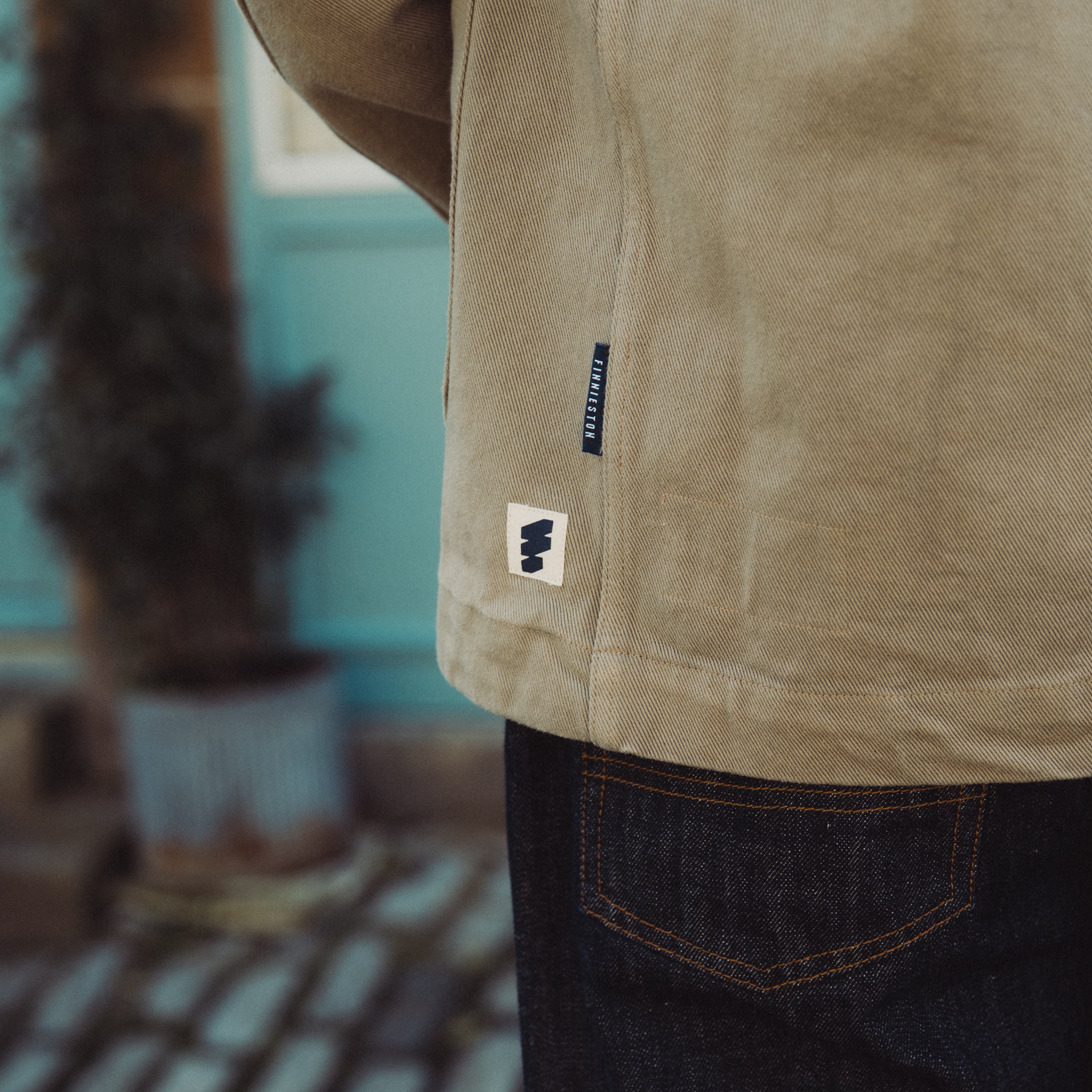Close-up of a person wearing a beige jacket with a brand logo, blurred indoor background