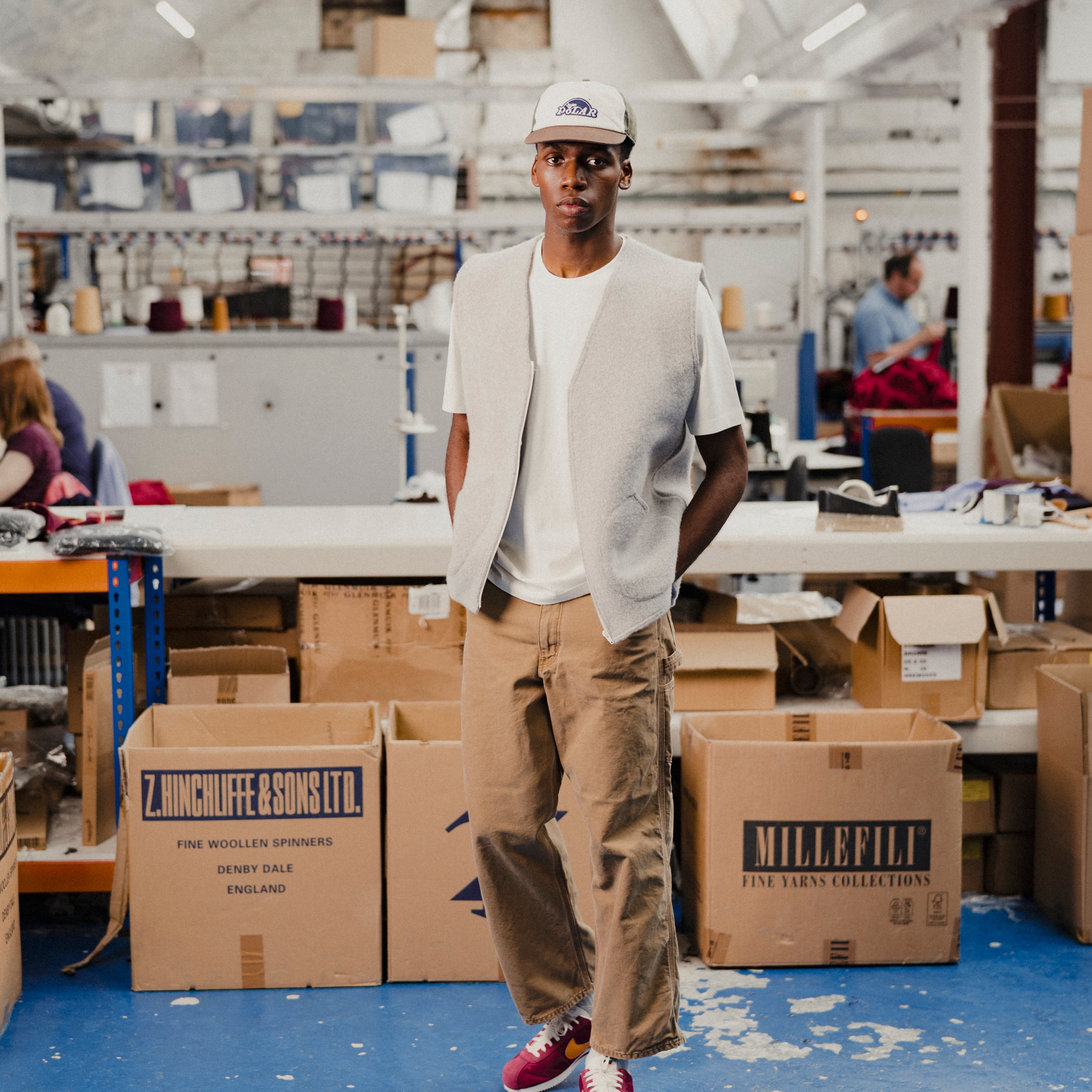 Person wearing a grey wool vest and white t-shirt standing in a warehouse with boxes and workstations in the background
