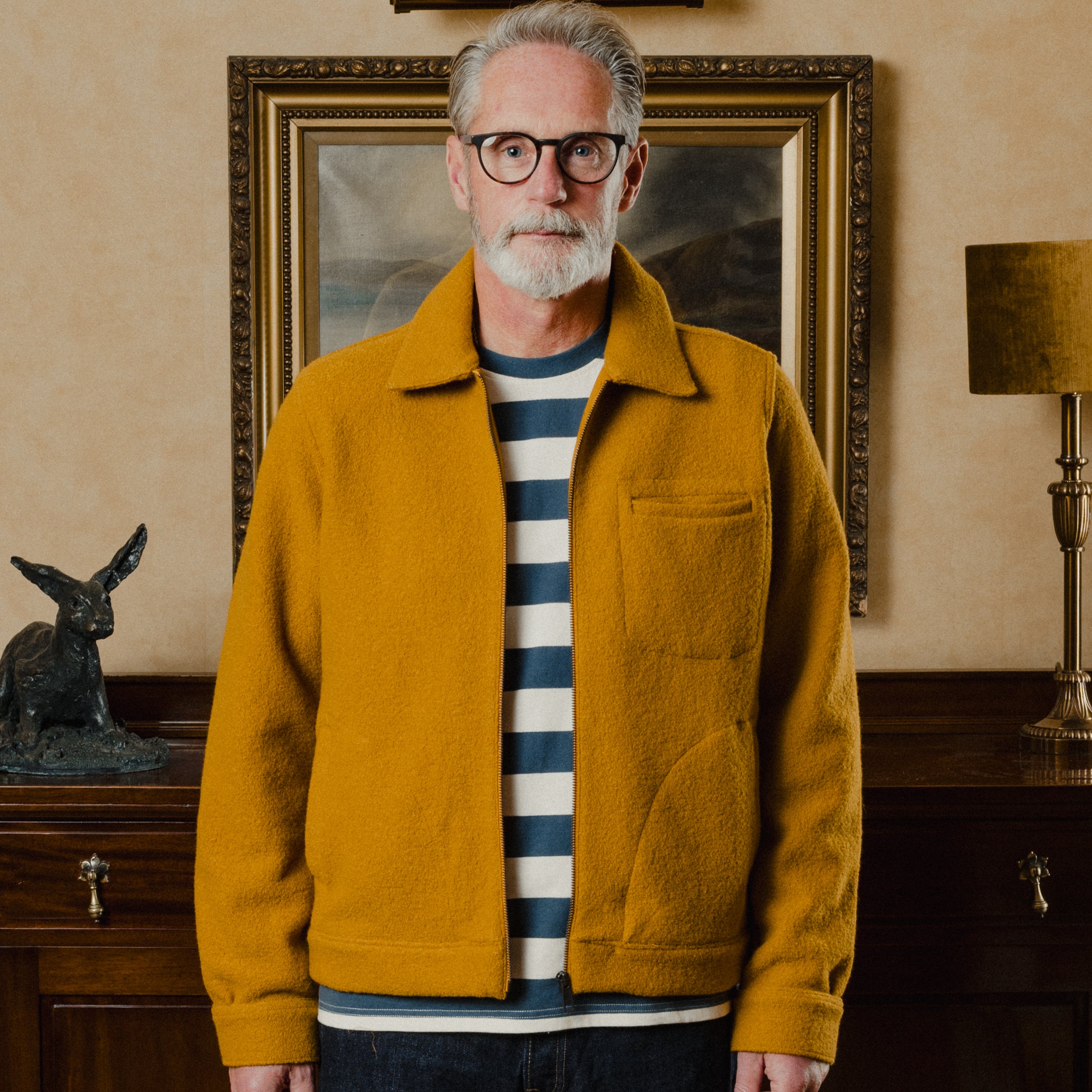 Man wearing a mustard yellow jacket in a room with a painting and lamp in the background