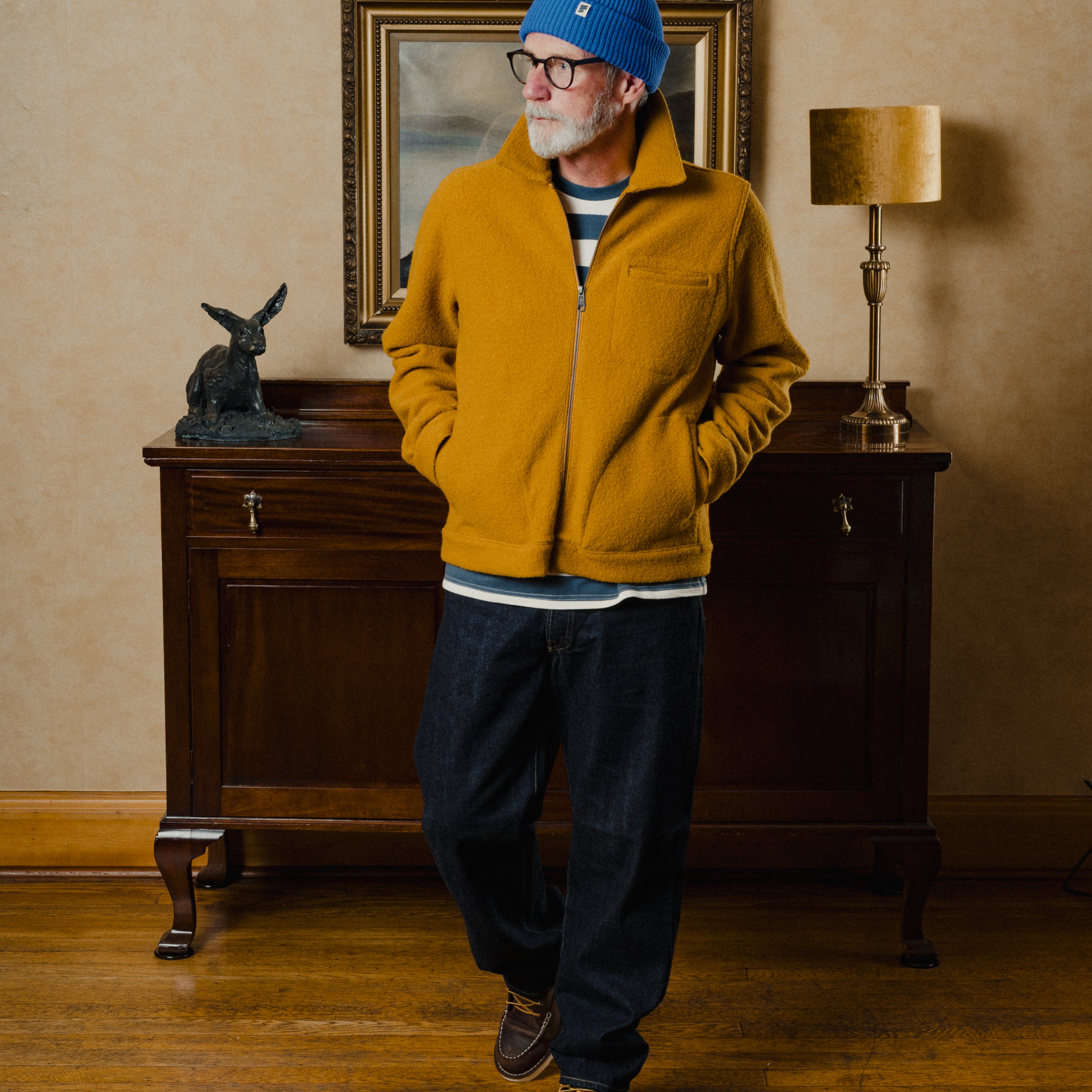Man wearing a mustard yellow fleece jacket and blue beanie standing in a room with wooden furniture and a lamp.