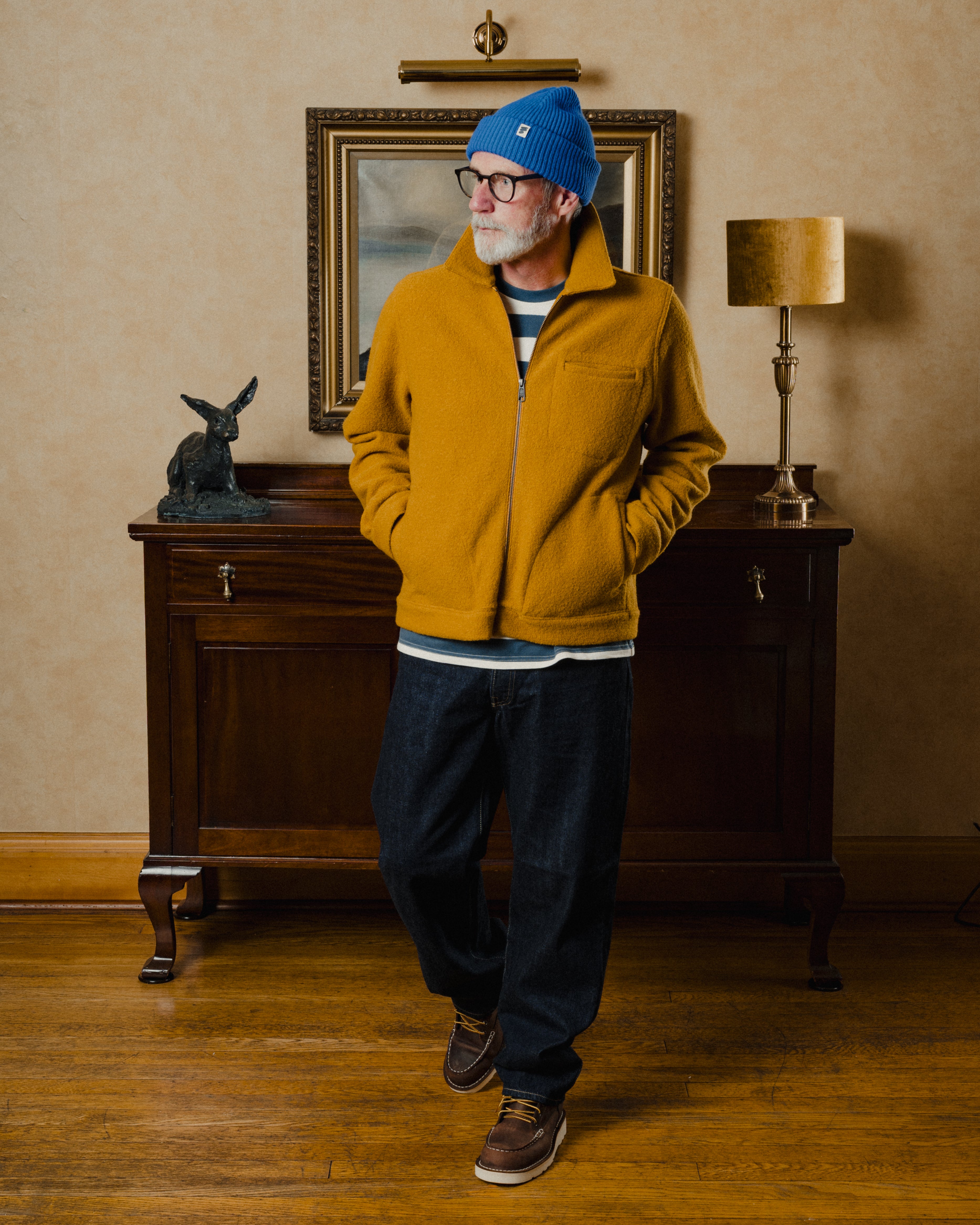 Man wearing a mustard yellow fleece jacket and blue beanie standing in a room with wooden furniture and a lamp.