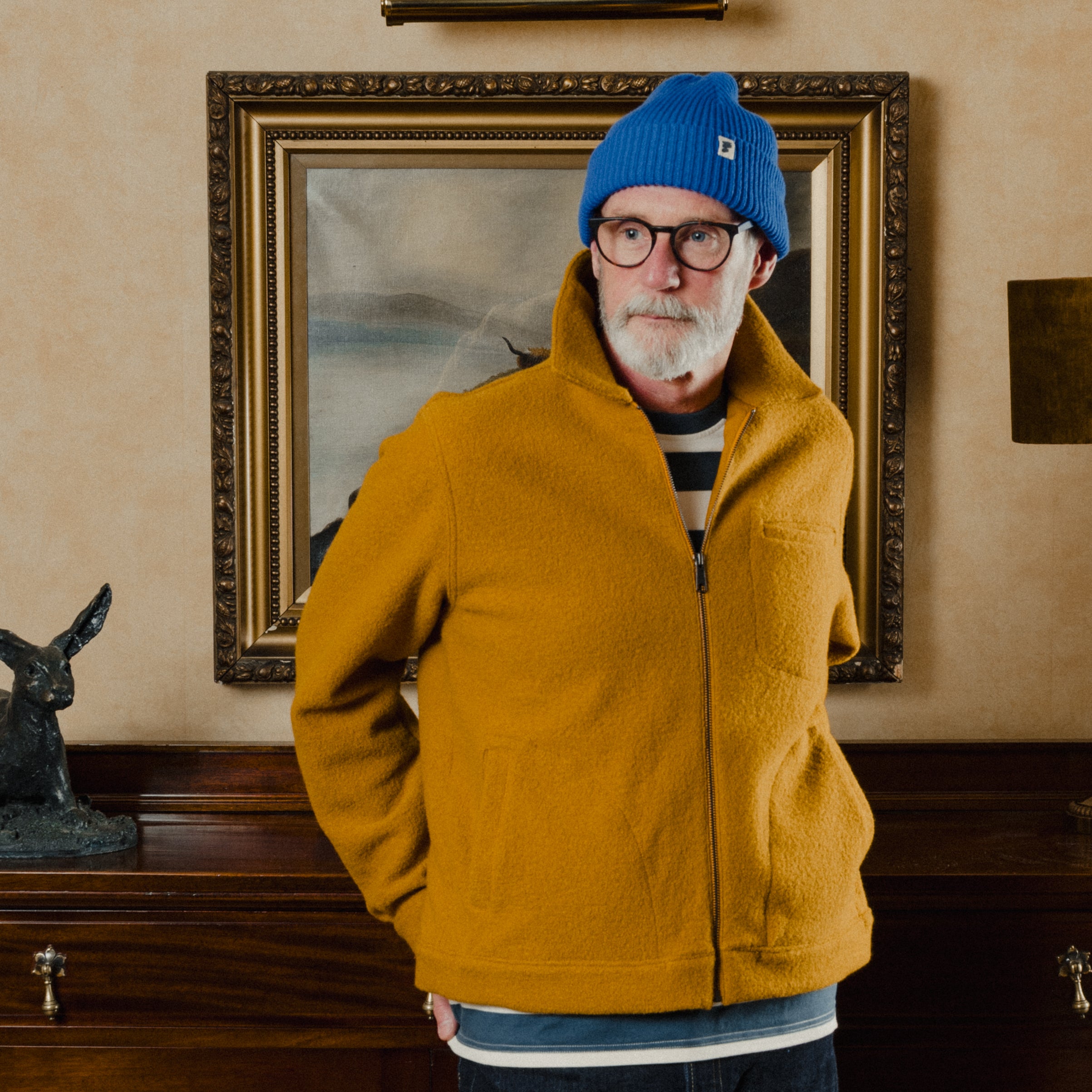 Man wearing a yellow jacket and blue beanie in a room with a painting on the wall.