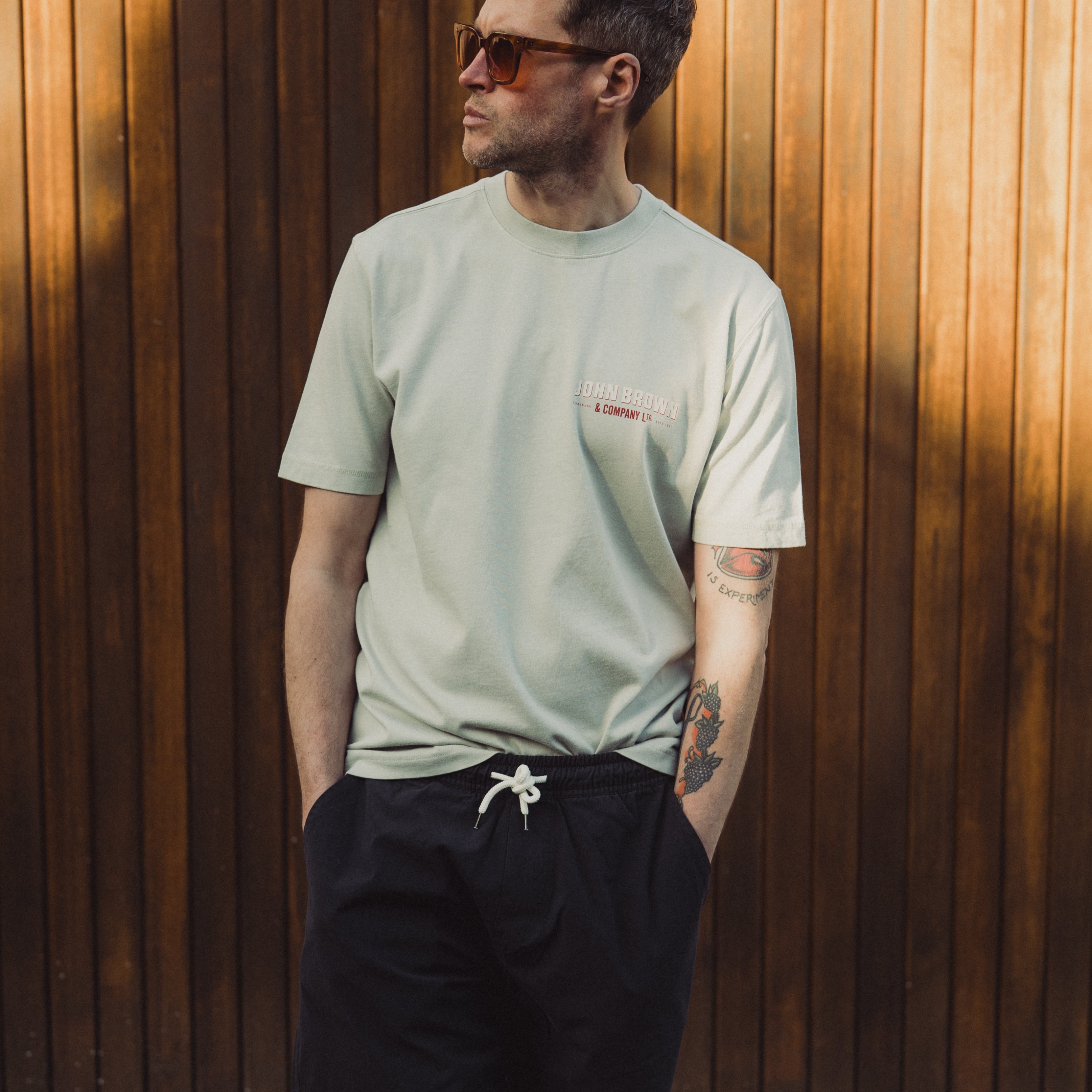 Man wearing a light green t-shirt and black shorts standing against a wooden wall.