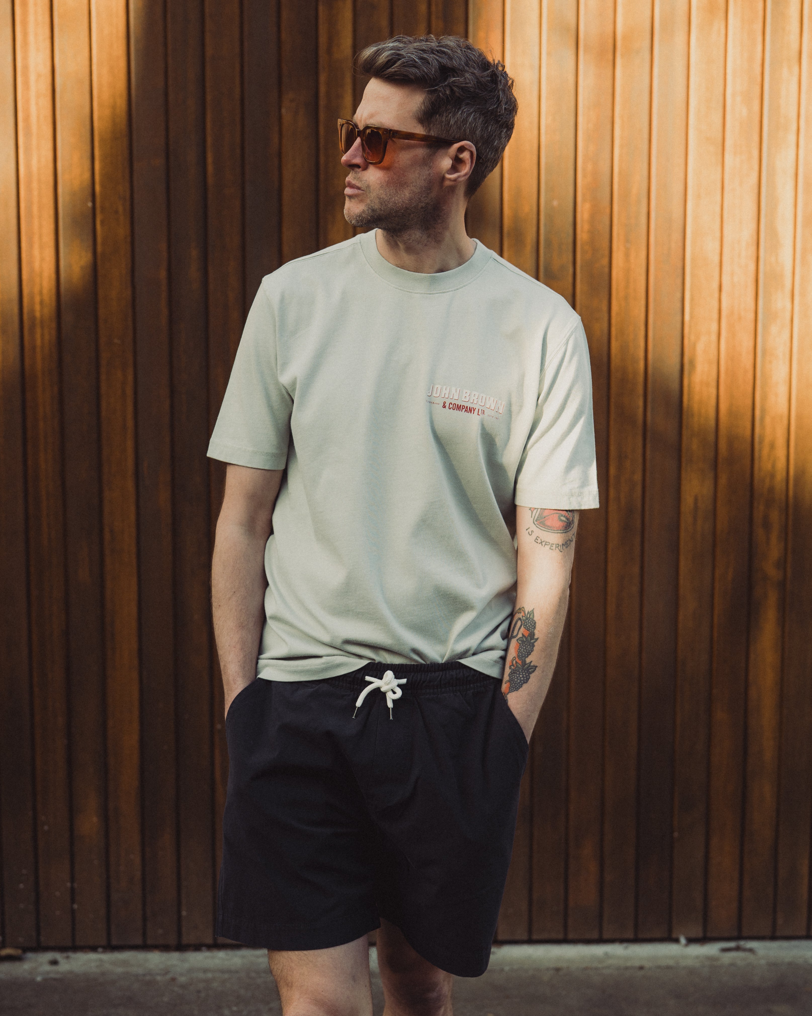 Man wearing a light green t-shirt and black shorts standing against a wooden wall.