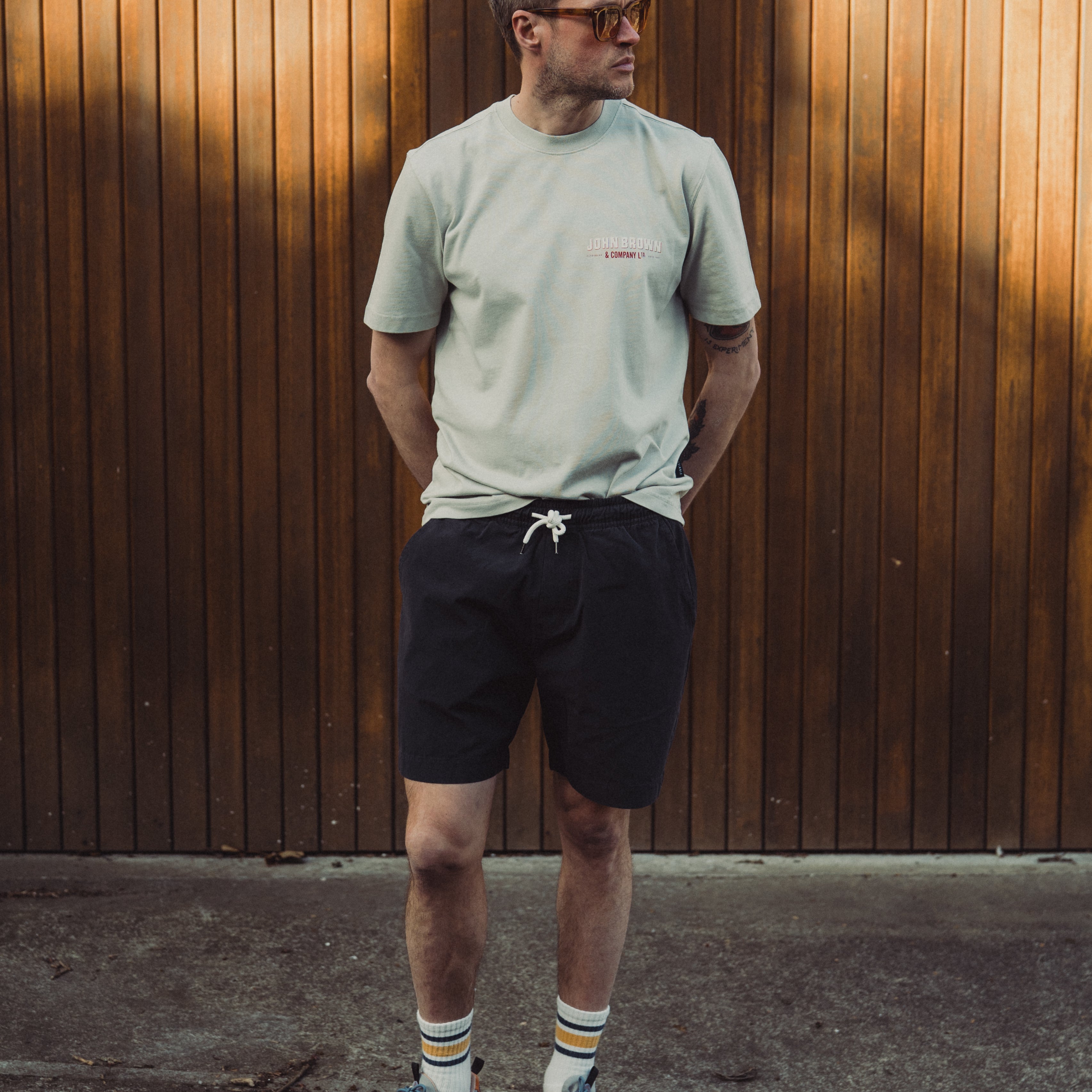 Man standing against a wooden wall wearing a light green t-shirt, black shorts, and colorful sneakers.