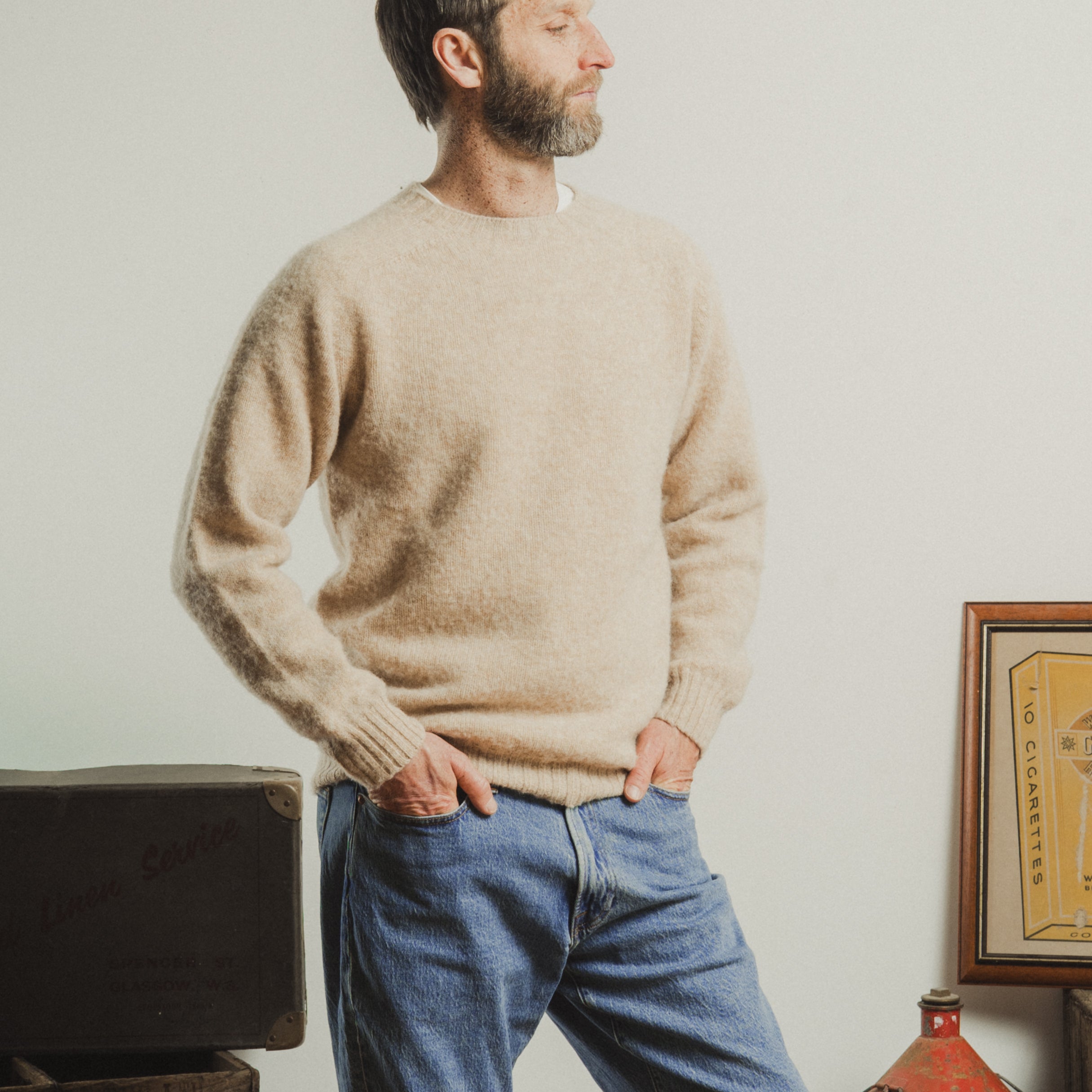 Man wearing a beige sweater and blue jeans standing in a room with a white wall.