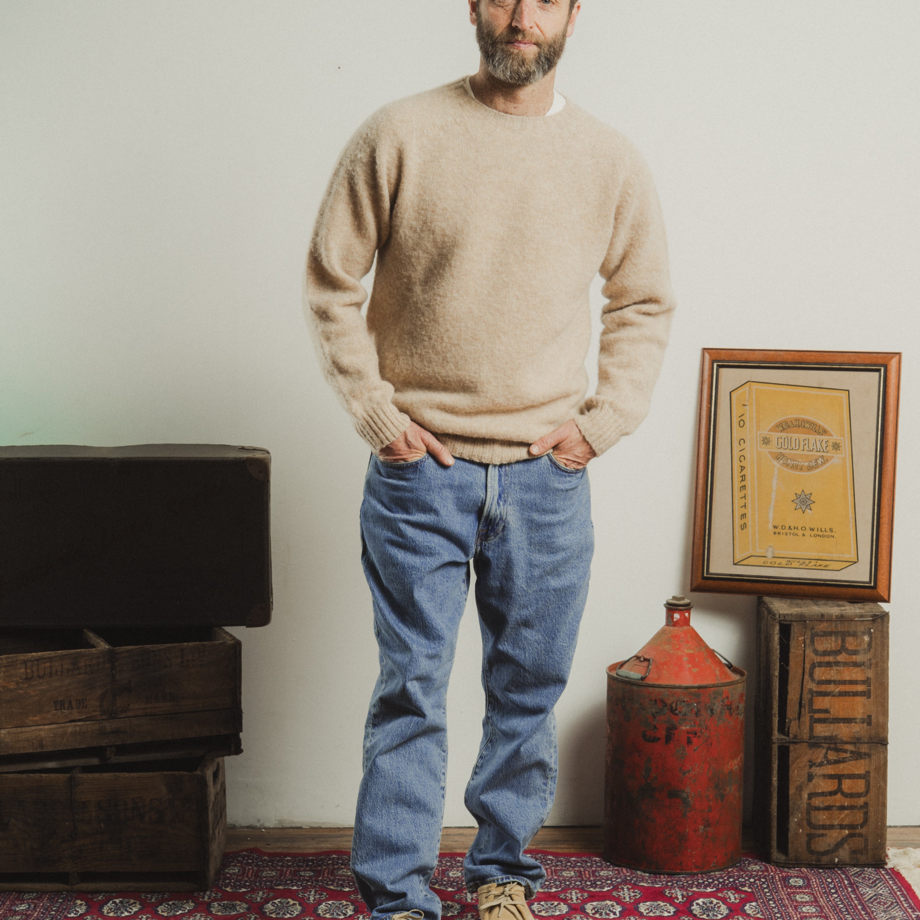 Man wearing a beige sweater and blue jeans standing in a room with a red rug and wooden crates.